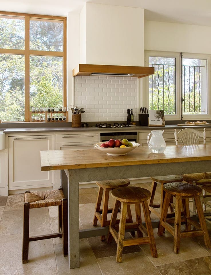 Une cuisine rustique avec une grande table en bois entourée de tabourets en bois. Il y a un bol de fruits et une carafe sur la table. La pièce comprend des armoires de couleur claire, un dosseret carrelé, de grandes fenêtres et une vue sur la verdure extérieure.