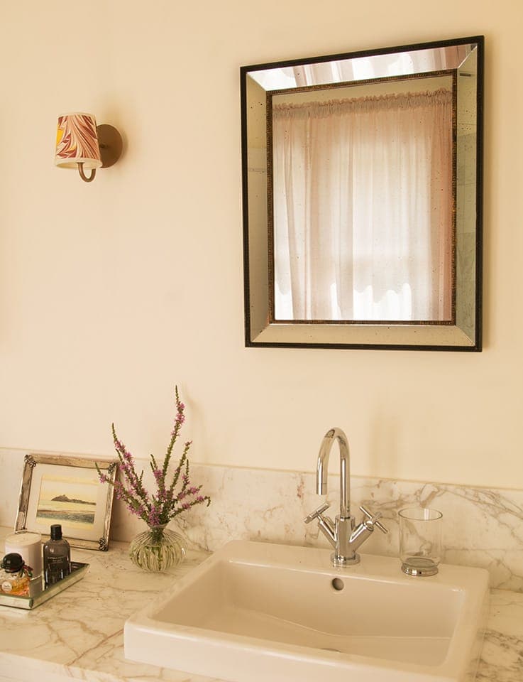Un comptoir de salle de bain avec un miroir rectangulaire au-dessus d'un lavabo blanc. Sur le comptoir en marbre se trouvent un tableau encadré, un vase avec des fleurs violettes, des produits de toilette et une applique murale avec un abat-jour à motifs. Un rideau transparent se reflète partiellement dans le miroir.
