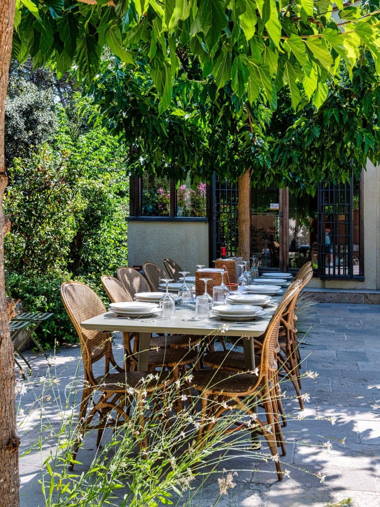 Une longue table à manger extérieure avec verres et assiettes est entourée de chaises en osier. L'espace est ombragé par des arbres verts feuillus et la lumière du soleil filtre à travers, créant un effet tacheté sur le sol pavé de pierres.