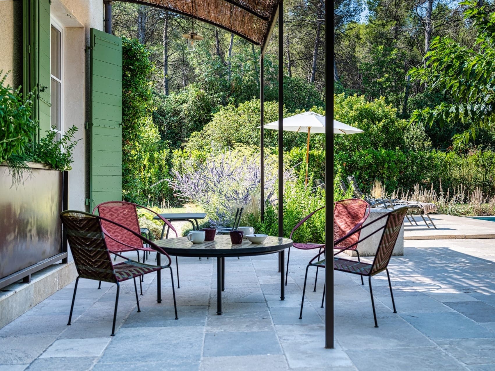 Un patio paisible avec une petite table ronde entourée de quatre chaises en osier. De la verdure, des arbres et un parasol blanc ornent l'arrière-plan, créant une ambiance extérieure sereine lors d'une journée ensoleillée.