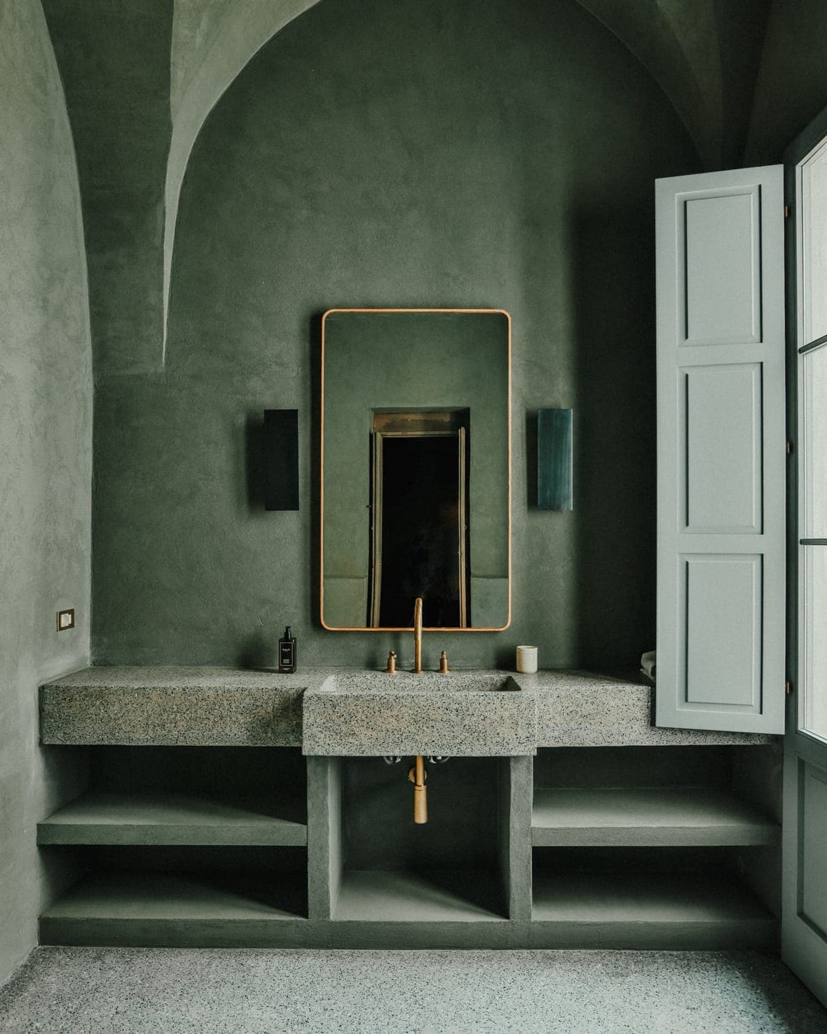 A minimalist bathroom features a concrete sink with an arched mirror framed in gold, flanked by modern sconces. Walls and shutters are painted in a muted green, creating a serene, elegant atmosphere.