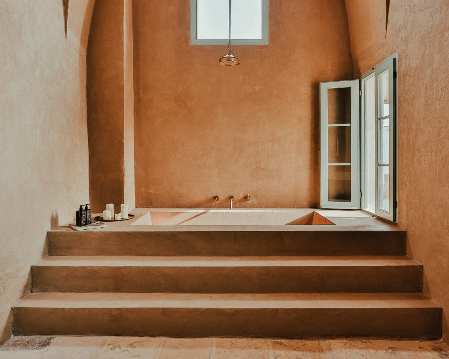 Minimalist bathroom with terracotta walls and sunken bathtub. Four wide steps lead up to the bath. A window and door open to the outside, allowing natural light to illuminate the space. Candles and bath products are placed nearby.