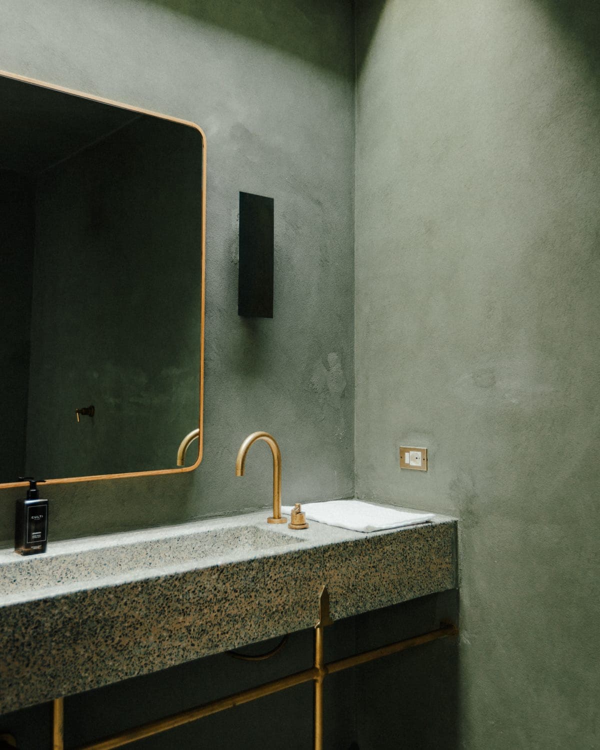 A minimalist bathroom features a textured concrete sink with brass faucet. A large mirror with a slim frame hangs above, and a black soap dispenser sits on the countertop. The walls are painted in a green hue, creating a serene atmosphere.