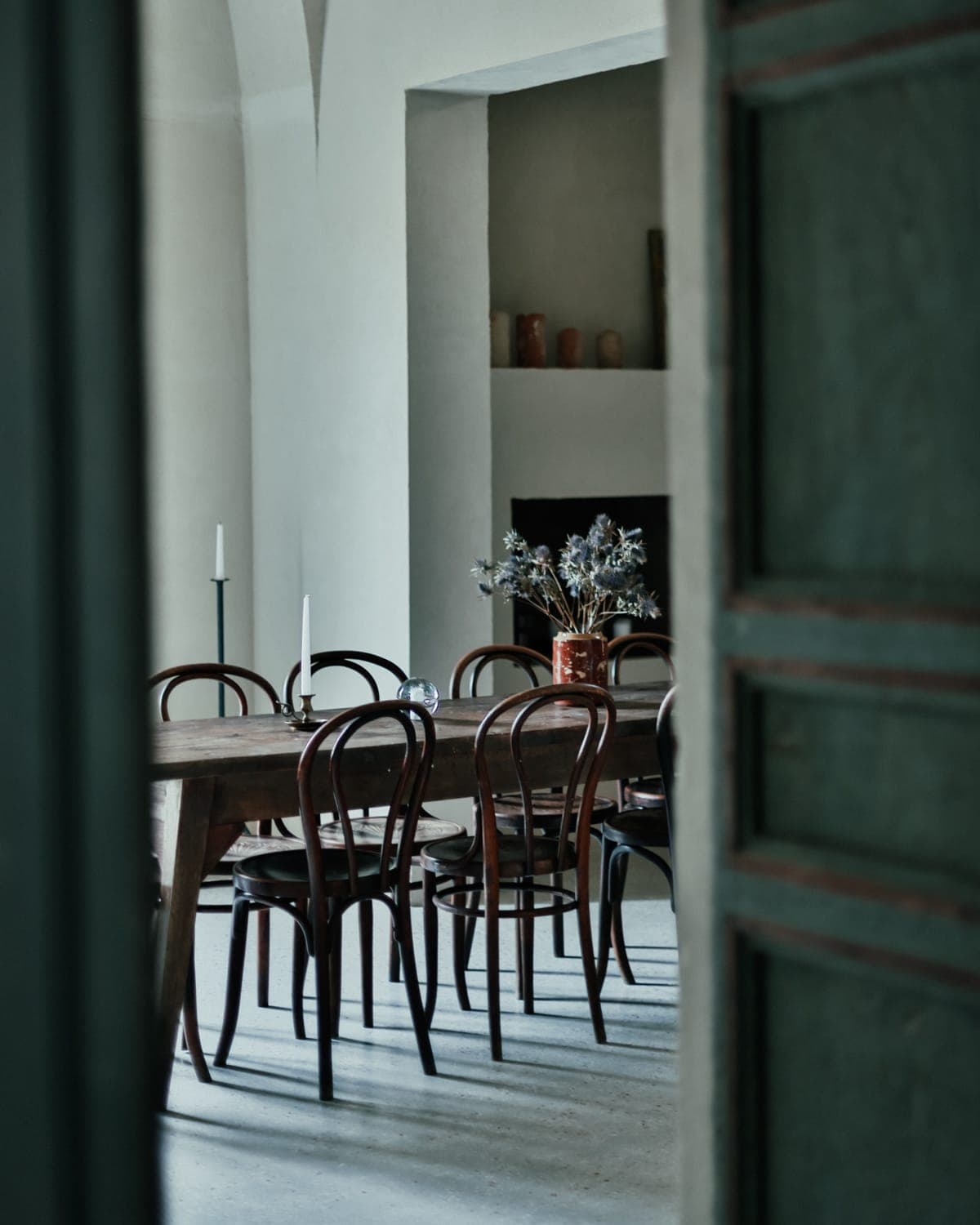 A softly lit dining room with a wooden table surrounded by several wooden chairs. The table is decorated with a vase of dried flowers and two candles. The room is visible through a partially open door, creating a warm, rustic atmosphere.