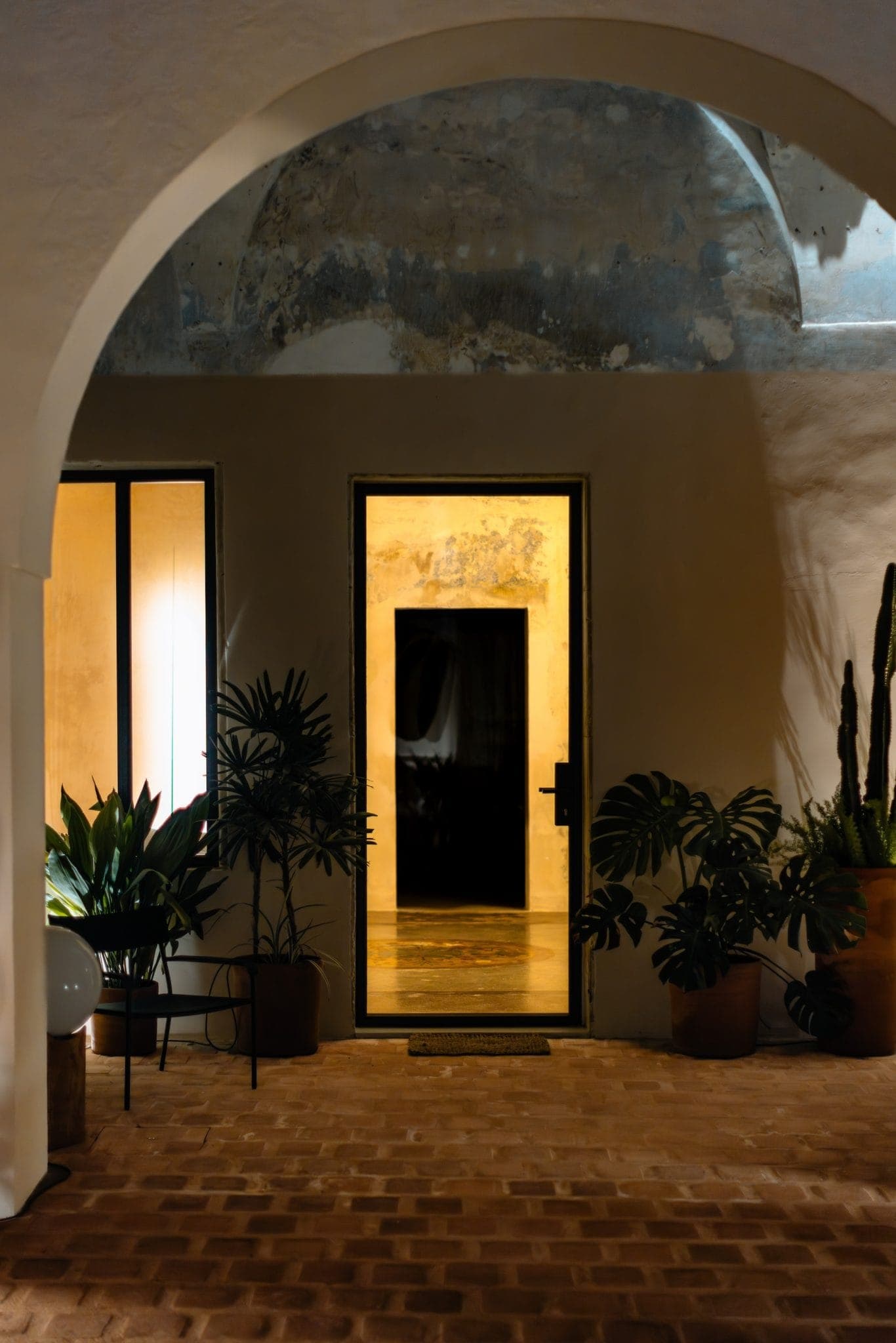 A warmly lit entrance with an open door under an archway at night. Potted plants and brick flooring create a cozy atmosphere.