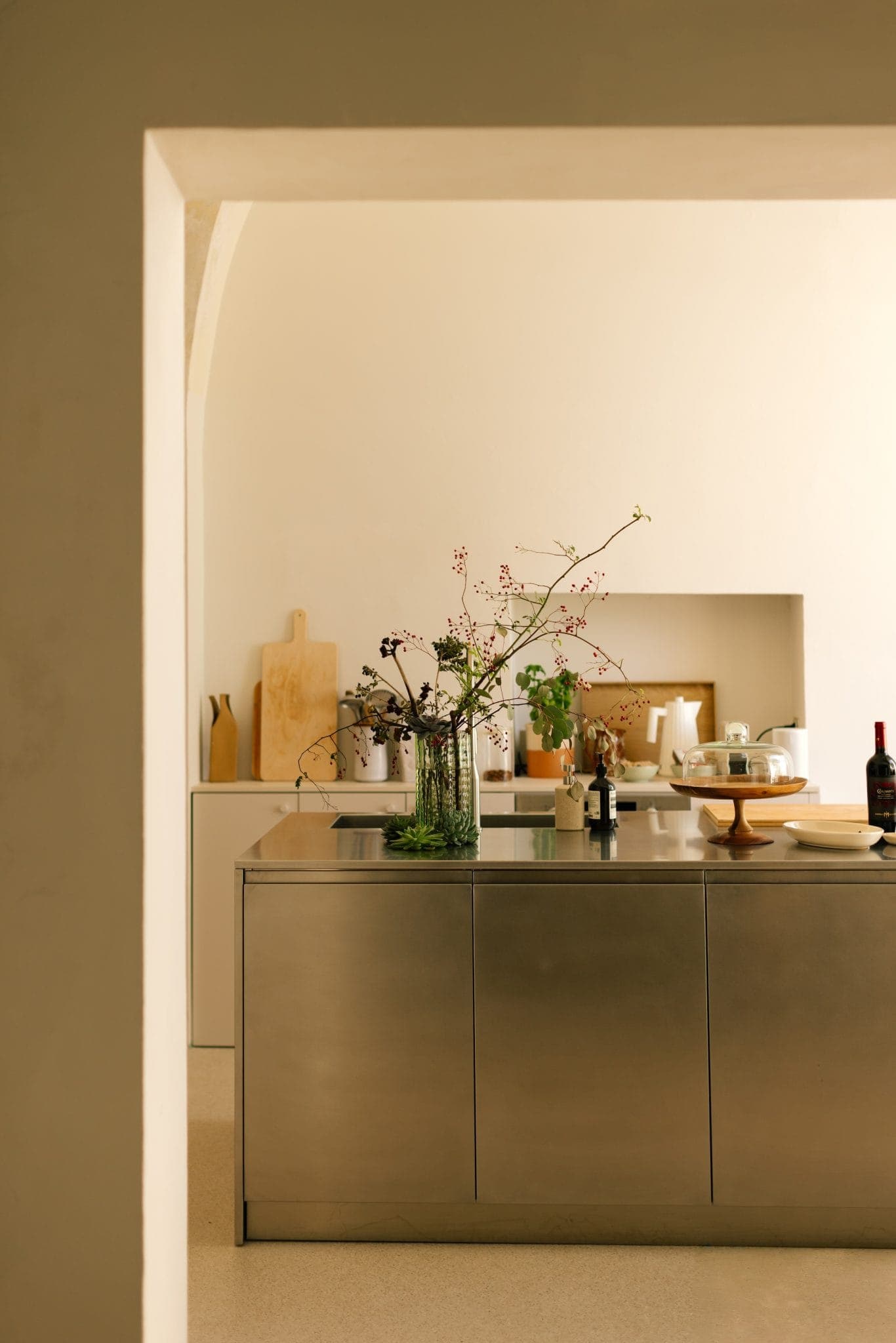 A minimalist kitchen with a sleek metal island featuring a vase of delicate flowers. The countertop holds cooking items and a wine bottle. The soft, neutral tones and natural light create a warm, inviting atmosphere.