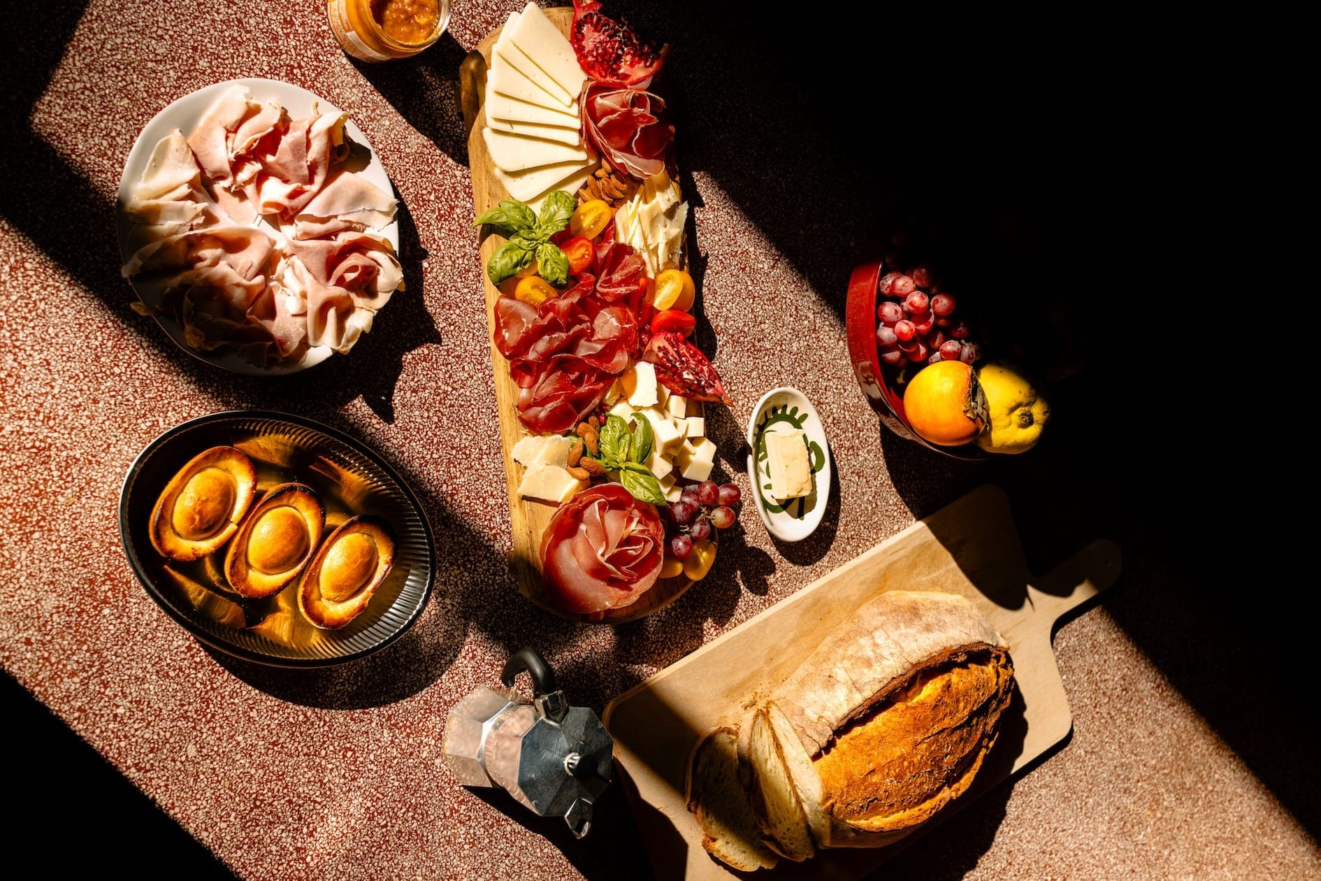A sunlit charcuterie board featuring sliced meats, various cheeses, grapes, and nuts. Nearby are a loaf of bread, roasted potatoes, butter, and assorted fruits. The arrangement is on a rustic tabletop with dramatic shadows.