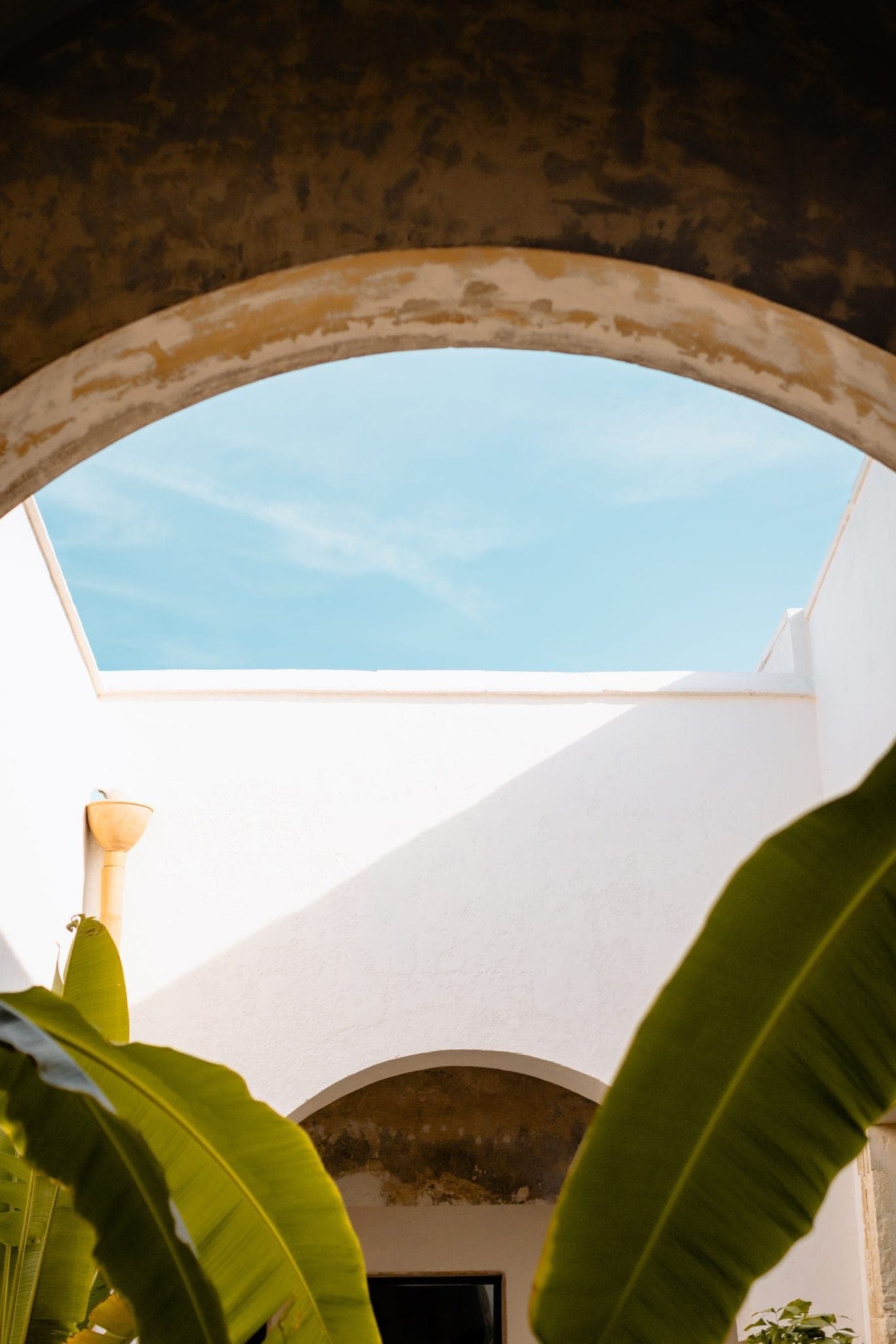 View through a large arched opening, showing a bright blue sky above white stucco walls. Large green banana leaves frame the foreground, adding a lush contrast to the scene.
