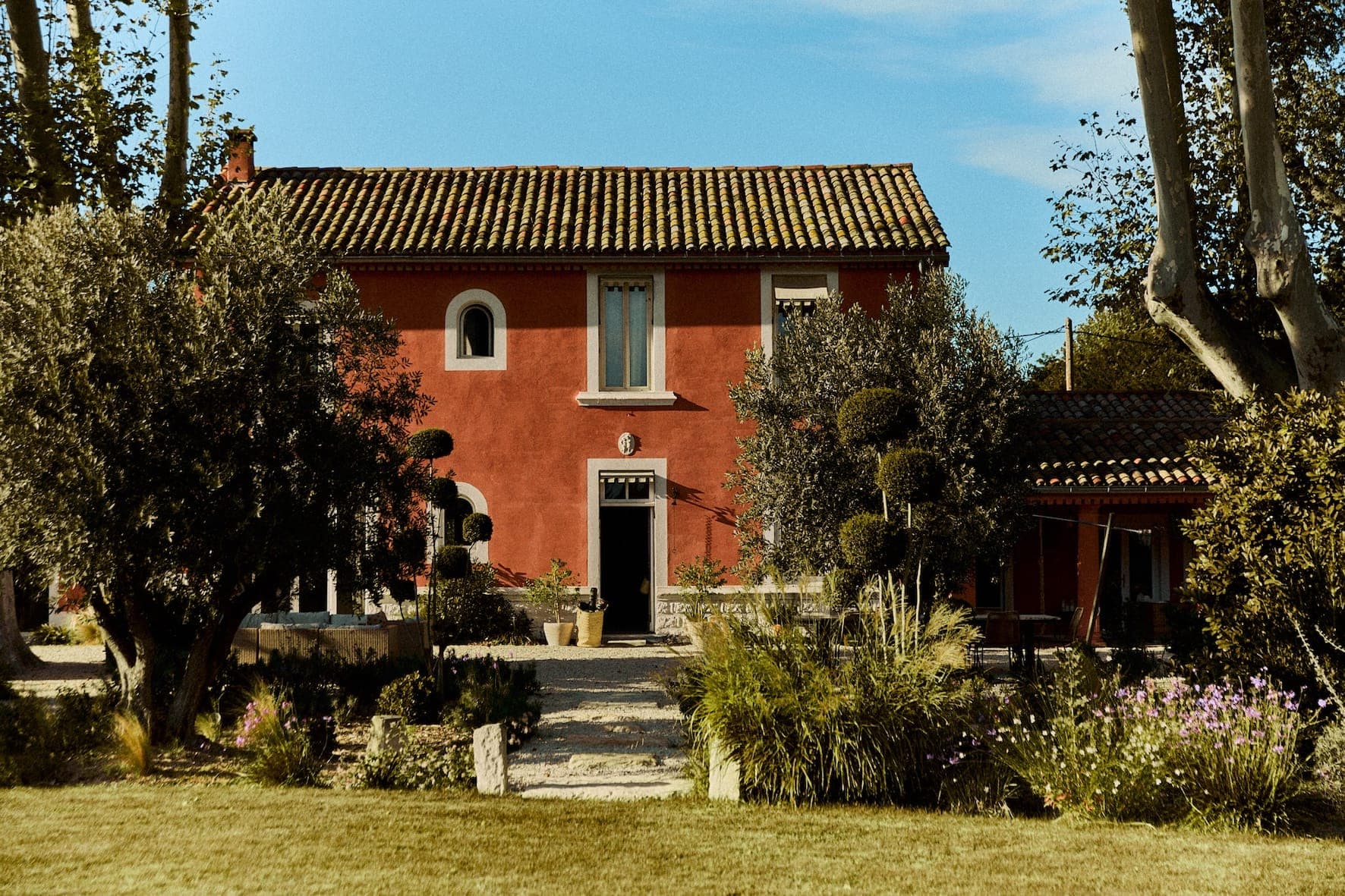Une maison rustique rouge avec un toit en tuiles, entourée d'une végétation luxuriante et d'arbres, et d'un jardin avec des fleurs. L'entrée comporte un chemin avec des plantes en pot, sous un ciel bleu avec des nuages épars.