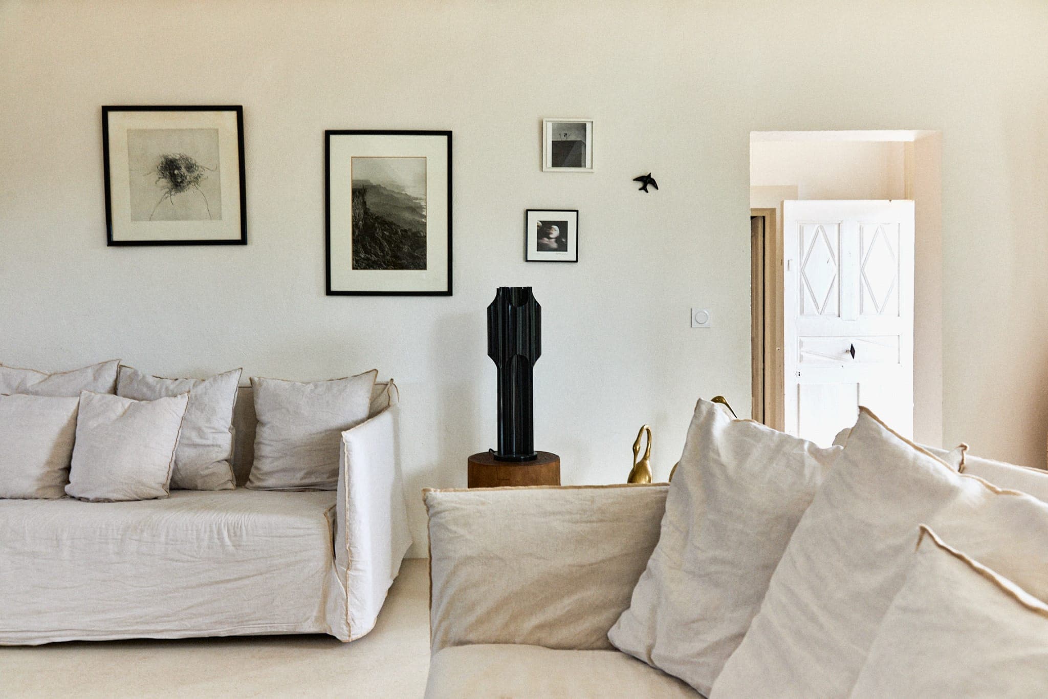 Un salon chaleureux avec des canapés aux couleurs claires et un mur blanc orné de tableaux encadrés. Une sculpture noire repose sur un socle en bois. La lumière du soleil filtre à travers une porte entrouverte, créant une ambiance chaleureuse.