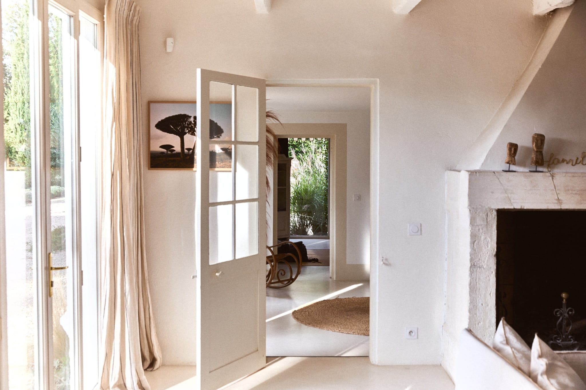 Une pièce lumineuse et aérée avec une porte ouverte menant à un couloir ensoleillé. La lumière naturelle traverse de grandes portes vitrées, illuminant une cheminée en pierre et un intérieur chaleureux et minimaliste. Une œuvre d'art encadrée en forme de silhouette et une chaise à bascule sont visibles.