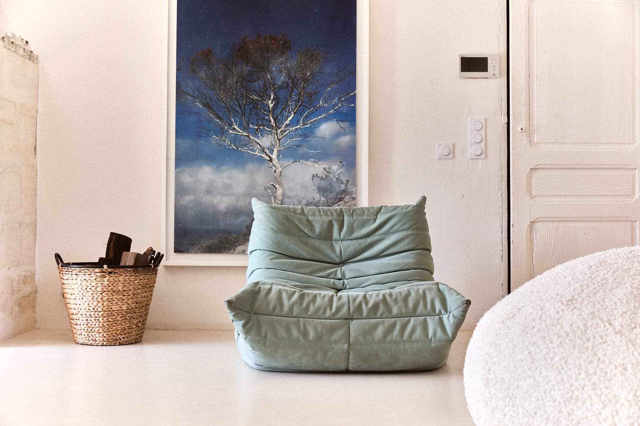 Une pièce confortable est dotée d'un fauteuil vert moelleux faisant face au spectateur. Une grande photographie d'un arbre sur fond de ciel bleu est accrochée au mur. Un panier tressé contenant des objets se trouve à proximité et une porte blanche est partiellement visible sur la droite.