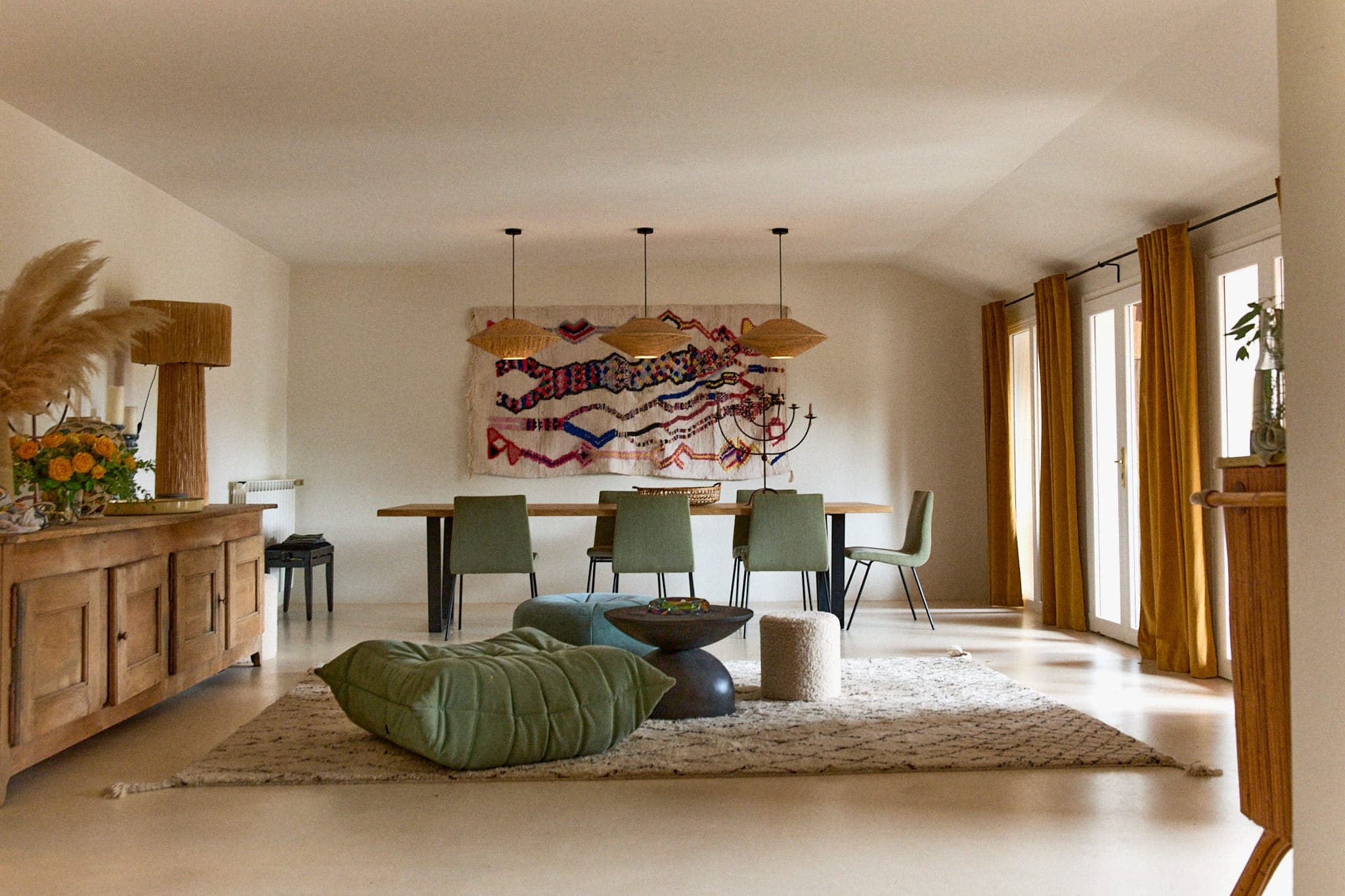 Une salle à manger confortable avec une longue table en bois et six chaises vertes. Une œuvre d'art textile colorée est accrochée au mur. Il y a des lampes suspendues au-dessus de la table et un coussin de sol vert moelleux sur un tapis beige. De grandes fenêtres avec des rideaux jaunes illuminent la pièce.