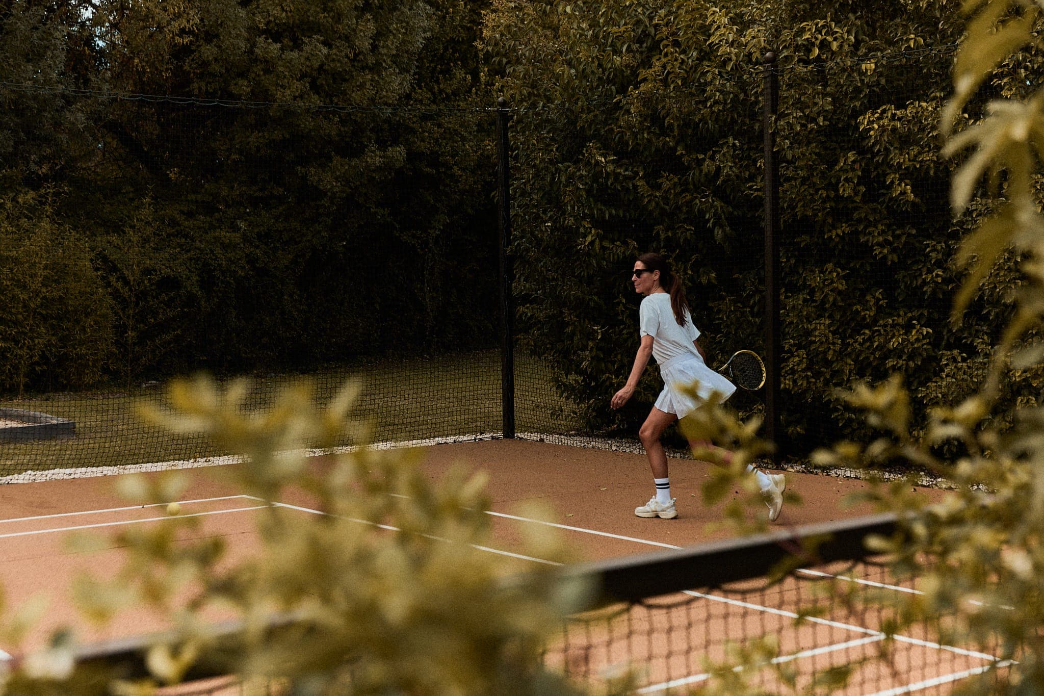 Une femme en tenue blanche joue au tennis sur un court en terre battue entouré de verdure. Elle s'apprête à frapper la balle, raquette en main et lunettes de soleil sur le nez. La scène est partiellement masquée par les plantes au premier plan.