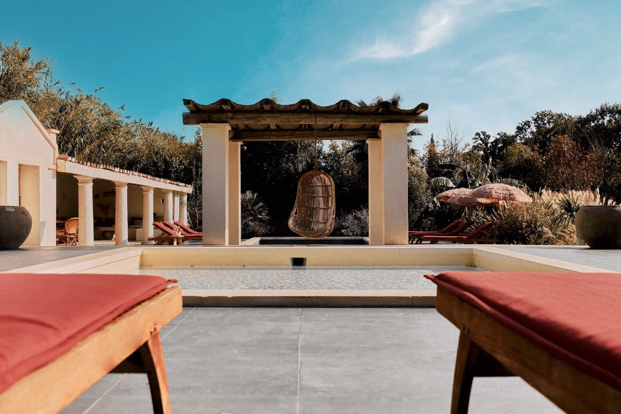 Des chaises longues face à une piscine tranquille, avec une chaise suspendue en osier sous une pergola. L'espace est entouré d'une végétation luxuriante et d'une structure extérieure. Le ciel est clair et bleu.