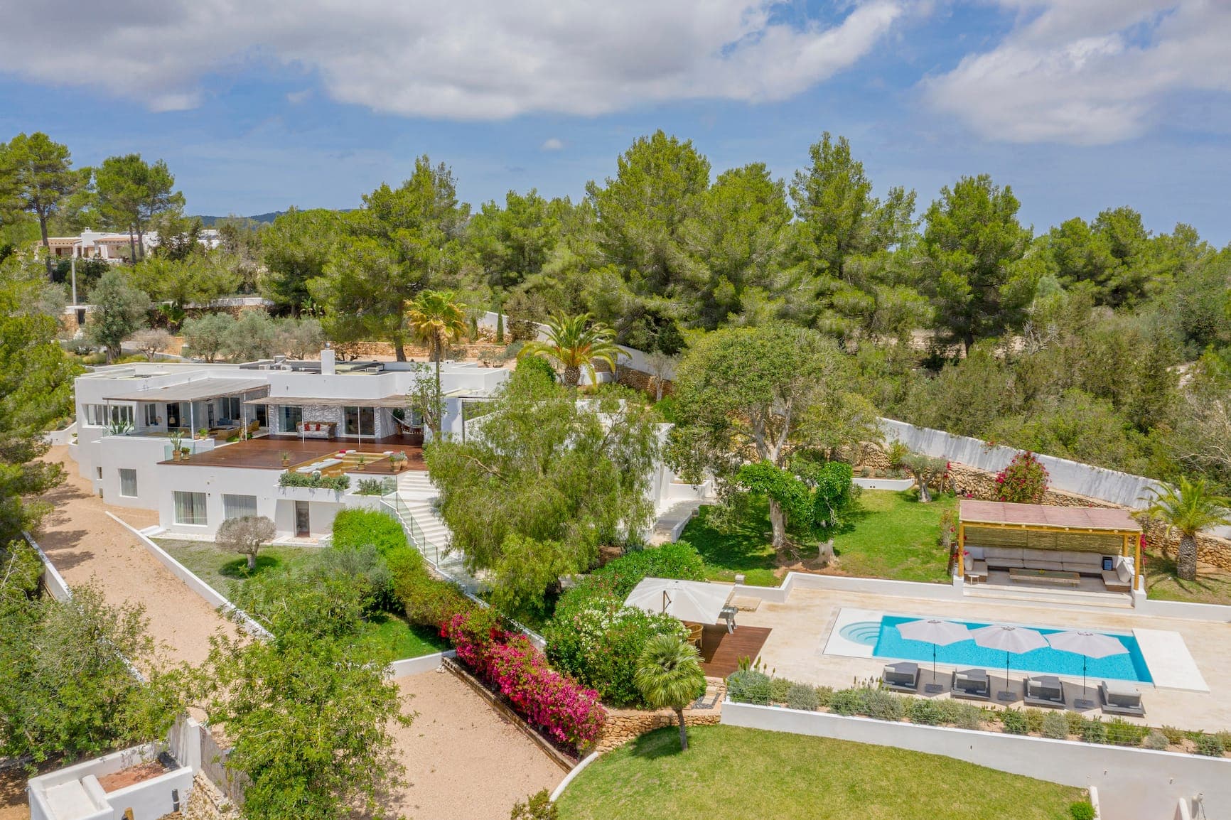 Vue aérienne d'une villa blanche moderne entourée d'une végétation luxuriante. La propriété dispose d'une piscine avec parasols bleus, d'une pergola en bois et de jardins paysagers. La villa dispose de plusieurs terrasses et se dresse sur un fond d'arbres et de ciel nuageux.
