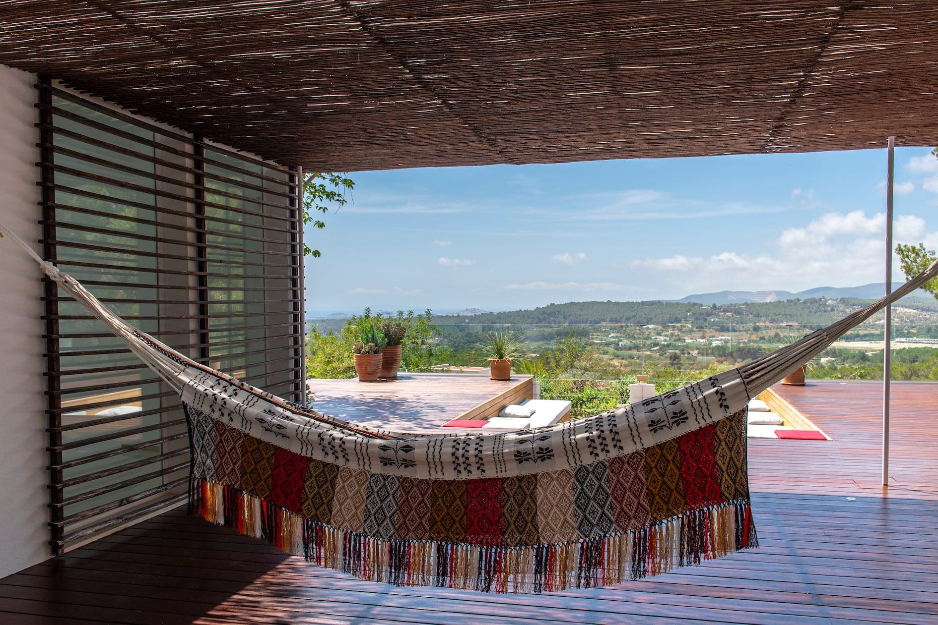 Un hamac coloré est suspendu dans une terrasse avec parquet, surplombant un paysage pittoresque avec des collines verdoyantes et un ciel bleu clair. Des plantes en pot et un banc en bois sont placés sur la terrasse.