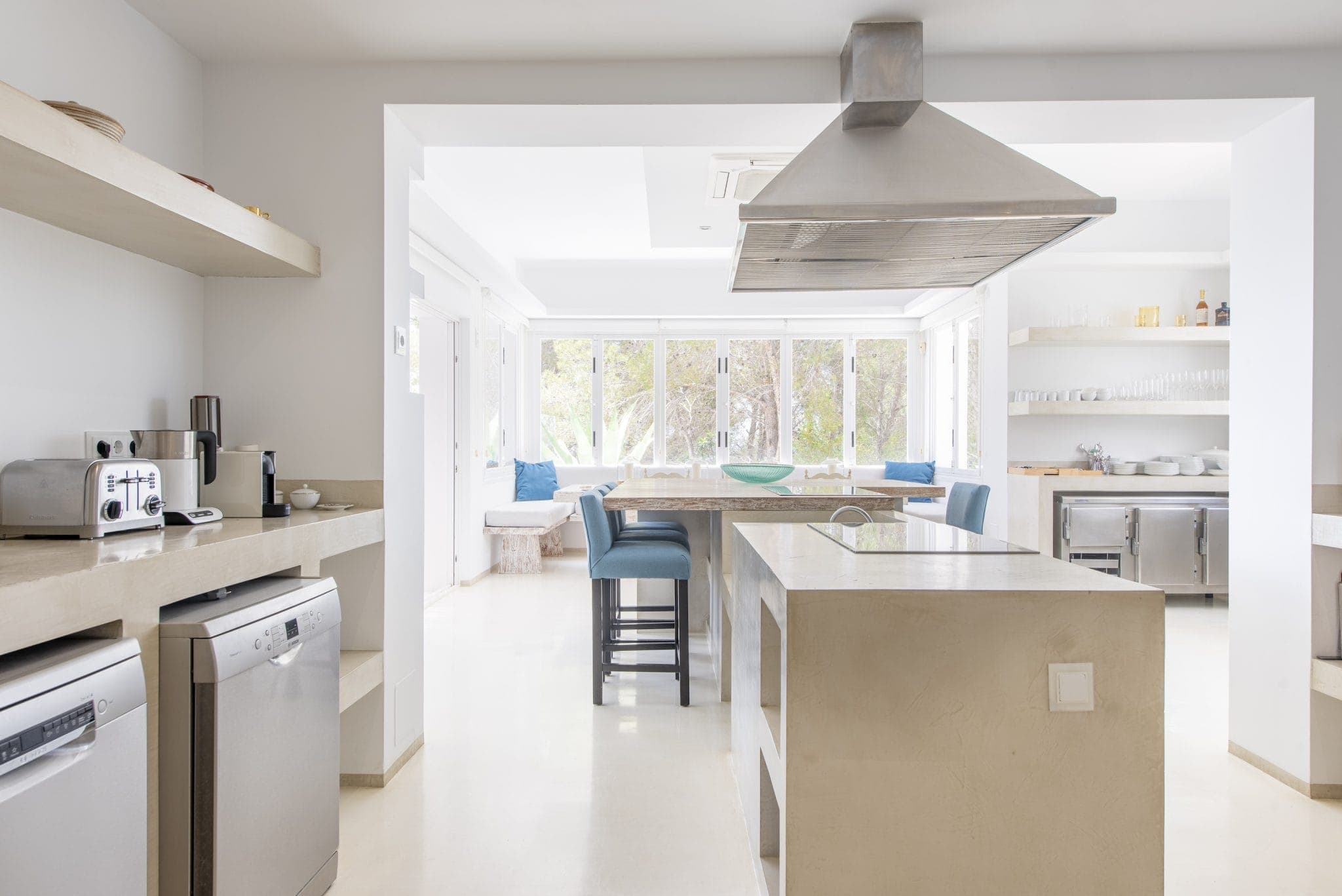 Cuisine moderne avec comptoirs de couleur claire et îlot. Elle comprend une hotte de cuisinière en acier inoxydable, un lave-vaisselle et un grille-pain. Il y a des étagères ouvertes avec de la vaisselle, une grande fenêtre avec vue sur le jardin et des tabourets bleus rembourrés au comptoir.