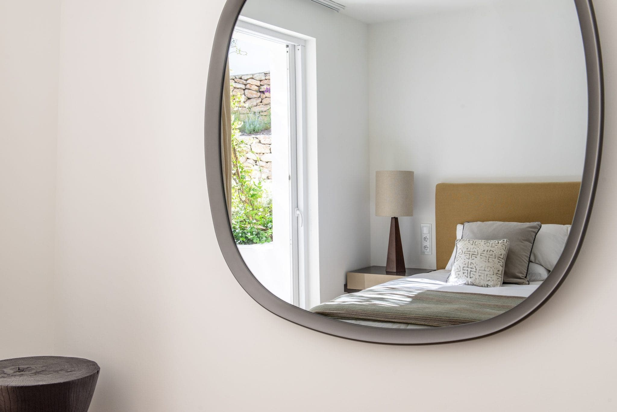 Une chambre moderne se reflète dans un miroir ovale sur un mur blanc. La pièce comprend un lit avec une tête de lit marron clair, un oreiller à motifs et une table de chevet avec une lampe. La lumière du soleil entre par une fenêtre, illuminant l'espace.
