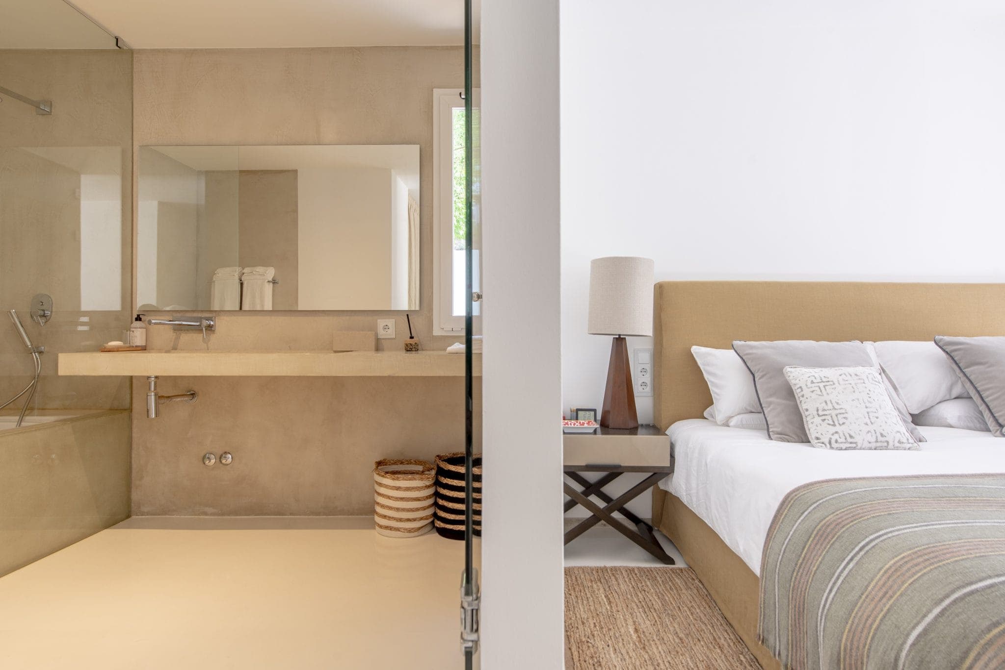 Un espace ouvert moderne comprenant une salle de bain et une chambre. La salle de bain comprend un lavabo beige, un grand miroir et une douche en verre. La chambre adjacente dispose d'un lit beige avec des oreillers blancs et gris, d'une table d'appoint et d'une lampe. Les tons neutres dominent la décoration.