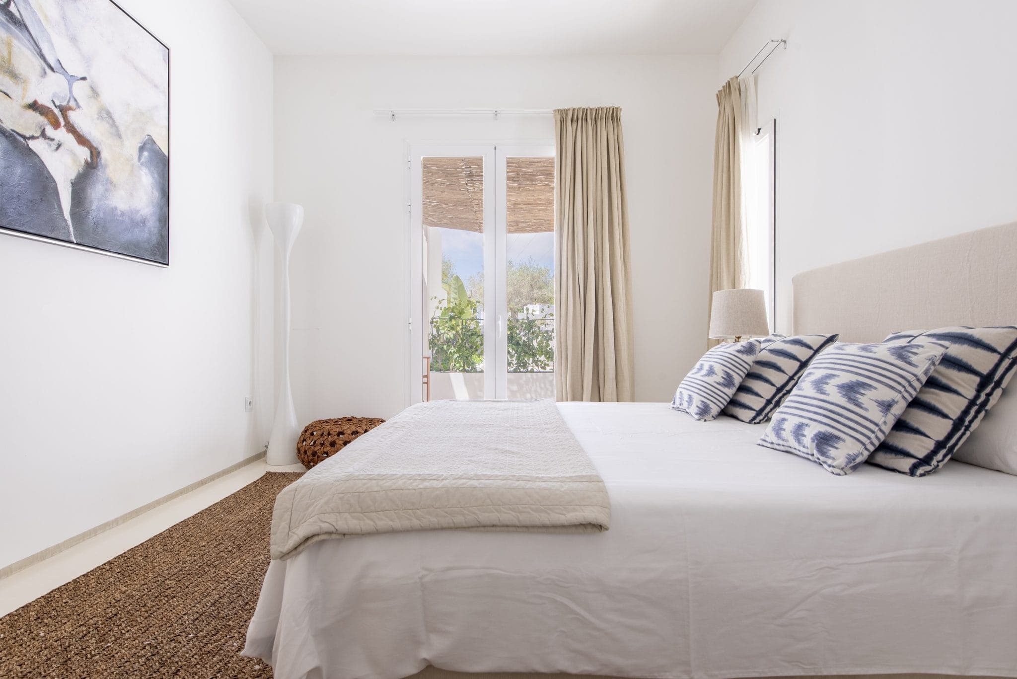 Une chambre lumineuse avec un grand lit orné de coussins à motifs et d'une couverture beige. Une grande lampe blanche trône dans un coin près d'un panier en osier. La chambre donne sur un balcon avec un arbre visible à travers les portes vitrées. Un tableau est accroché au mur.