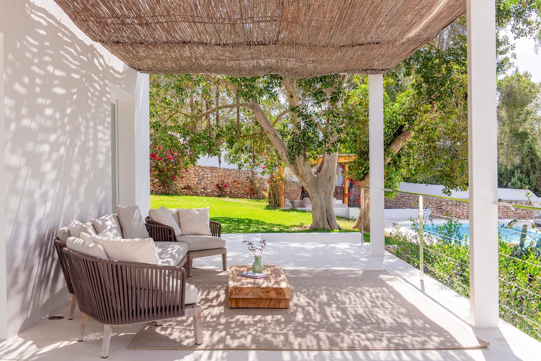 Un patio confortable avec deux chaises en osier et une petite table en bois sur un tapis clair. L'espace est ombragé par un toit tressé, donnant sur une pelouse verte luxuriante et un grand arbre. En arrière-plan, il y a un mur de pierre, un jardin et un aperçu d'une piscine.