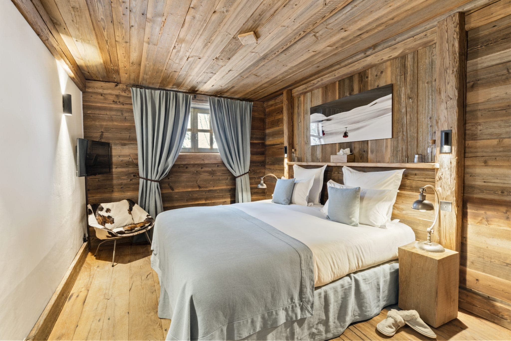 Comfortable wooden bedroom with a double bed covered in light blue linen. A cowhide chair sits in the corner next to a small wooden side table. Curtains frame a window and a flat-screen TV is fixed to the wooden wall. A mirror hangs over the bed.