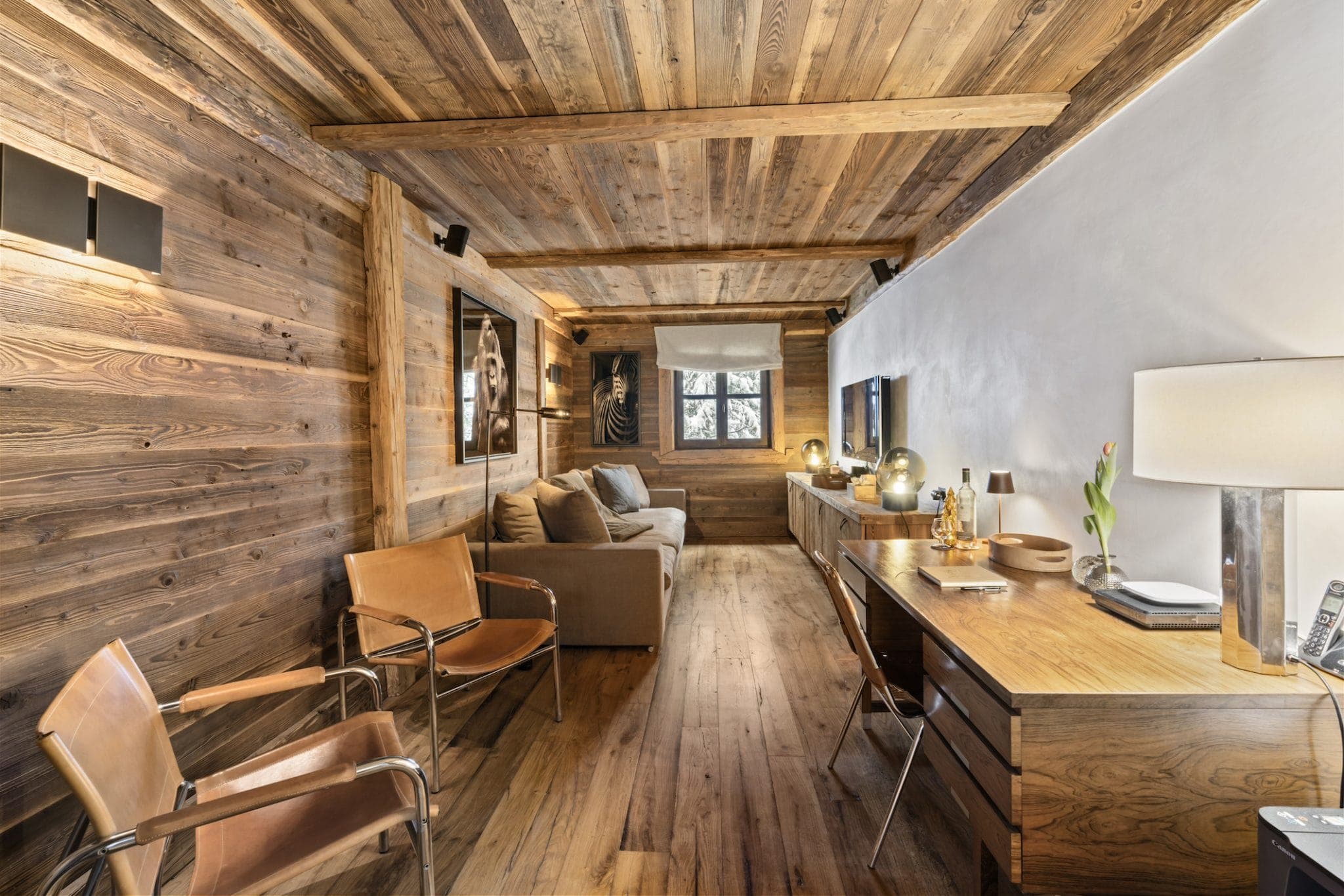 A comfortable, narrow room with wooden walls and ceiling, with a long desk on the right, a sofa at the end and two leather chairs on the left. The room is softly lit by lamps and has a small window offering natural light.