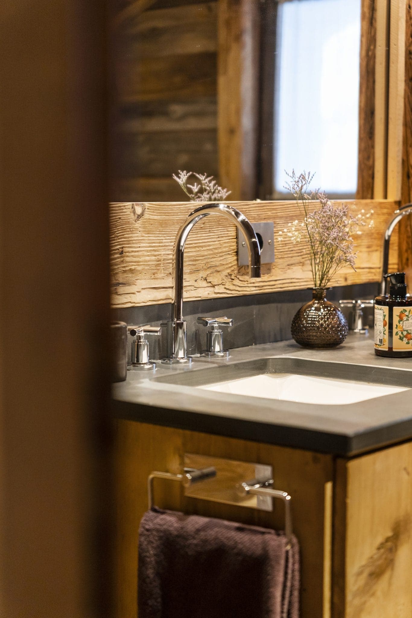 A rustic sink with an elegant metal faucet is surrounded by a wooden worktop and wall. A small vase with dried flowers and a bottle of liquid soap sit on the countertop. A dark towel hangs from a rack beneath the sink.