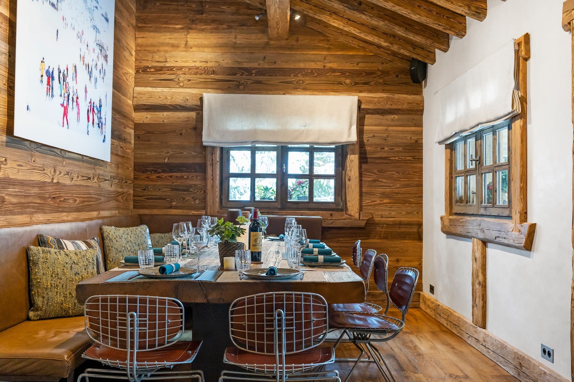 A comfortable dining room with a wooden interior, including a long table set with plates, glasses and napkins. The room has a large window, a bench on one side and several chairs. A work of art depicting a snowy landscape hangs on the wall.