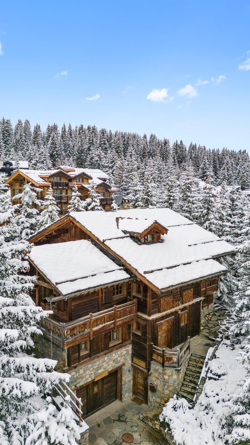 A large wooden chalet with a snow-covered roof is nestled among snow-laden pine trees. The scene is bright with a clear blue sky, and other similar structures are visible in the background, creating a serene winter landscape.