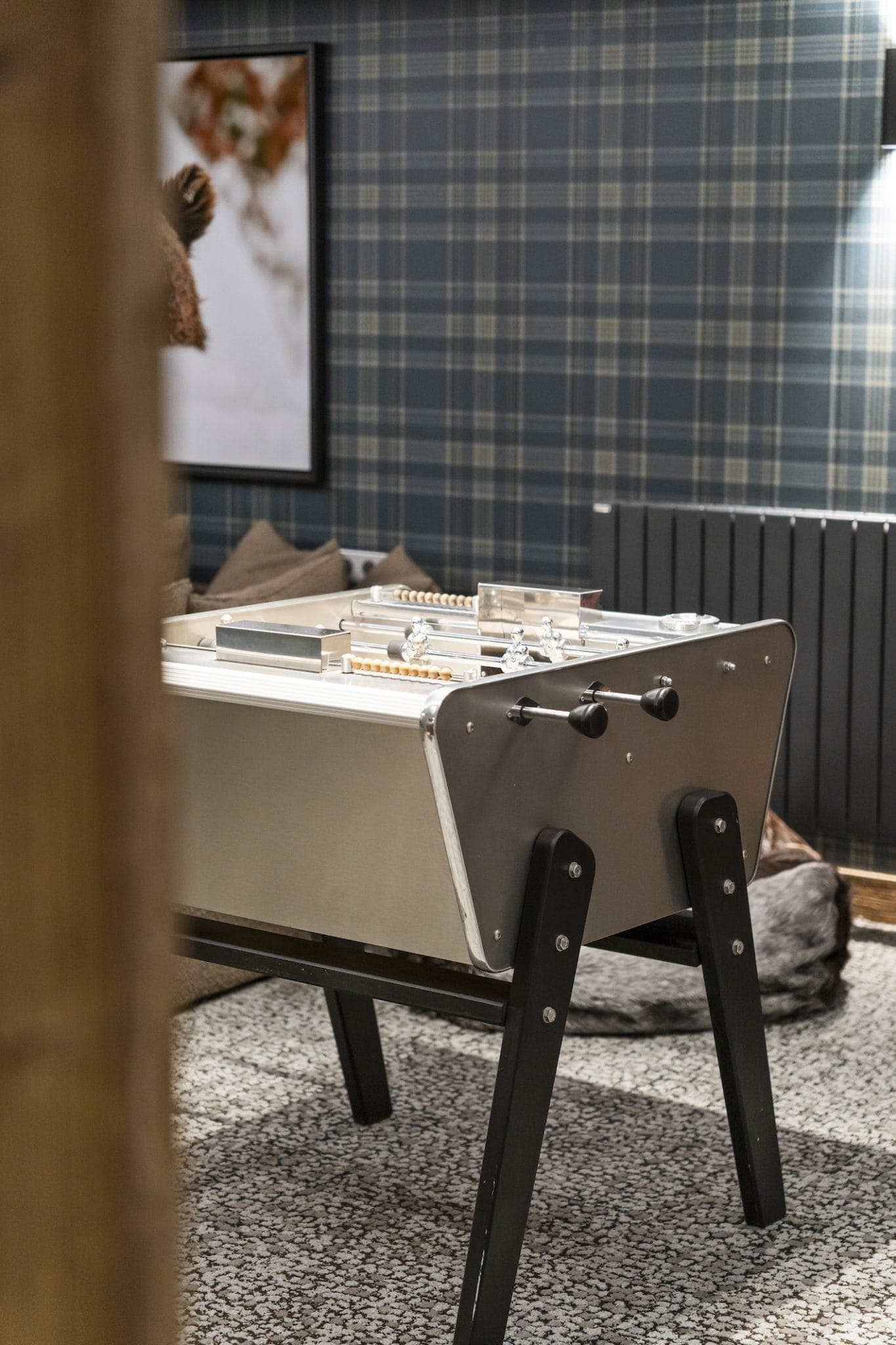 A foosball table with black legs and a metal playing surface is set in a room with checkered wallpaper and textured carpet. A framed painting and a radiator are visible in the background.