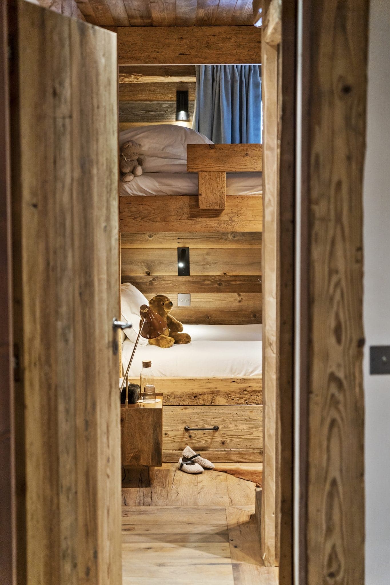 The picture shows a comfortable wooden bedroom with two bunk beds. Each bed has white bedding and a teddy bear. Soft lighting illuminates the space, and slippers are placed on the floor next to the bed. A small glass of water is placed on the bedside table.