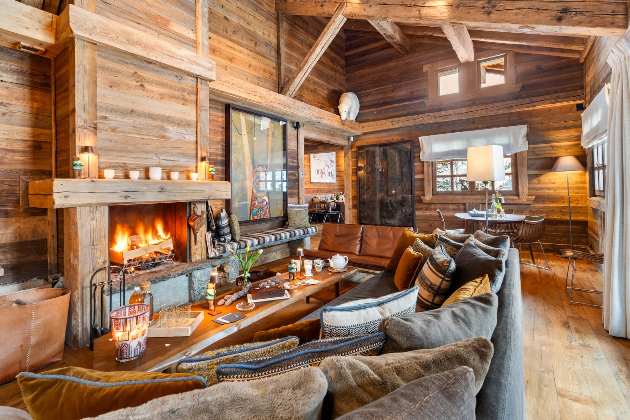 Cozy rustic living room in Chalet IMA with wooden walls and beams, featuring a lit fireplace. A large L-shaped sofa with cushions faces the fire. Candles and decor items adorn the space. Natural light enters through windows, enhancing the warm atmosphere.