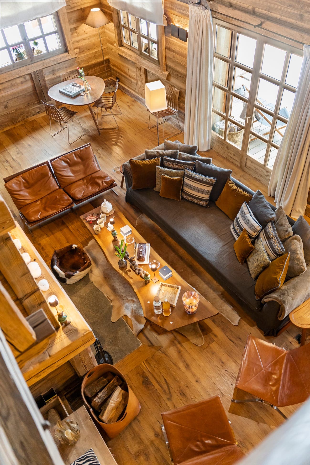 Warm, rustic living room with wooden walls and floors, large sofa with cushions and long coffee table. Leather chairs and a cowhide rug complete the decor. A round dining table is set in the corner, under a window with white curtains.