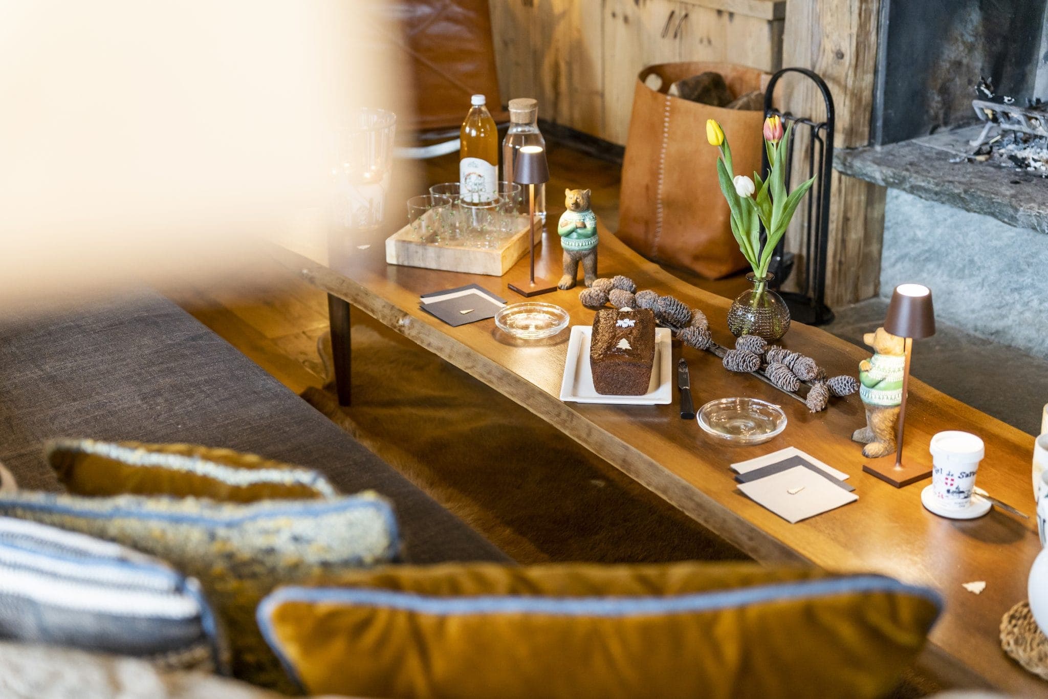 A cozy living room scene with a wooden coffee table holding a cake, pinecones, tulips in a vase, and various decorative items. Soft cushions are on the sofa and there's a bag and fireplace in the background, creating a warm, inviting atmosphere.