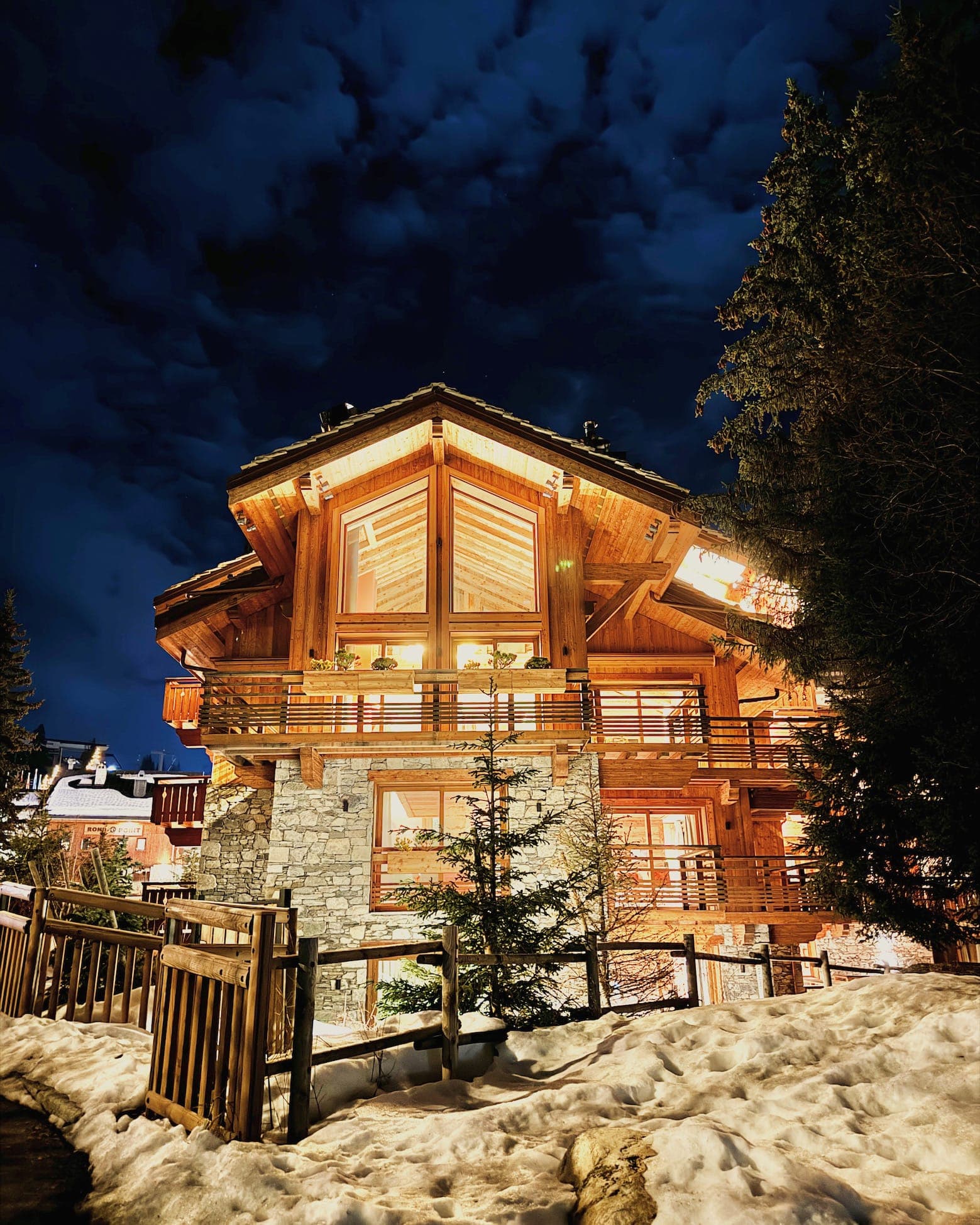A warmly lit wooden chalet with stone accents, surrounded by snow and a wooden fence, under a cloudy night sky. Evergreen trees are nearby, adding to the cozy, wintery atmosphere.