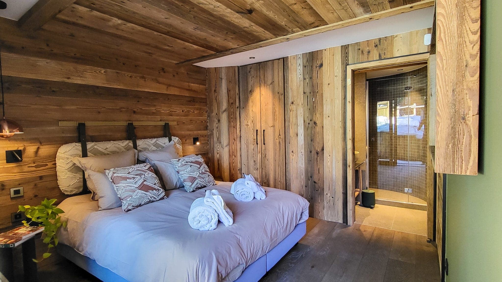 Cozy bedroom with rustic wooden walls and ceiling, a comfortable double bed adorned with decorative pillows and neatly folded towels. A doorway leads to a modern, tiled bathroom. Warm ambient lighting enhances the inviting atmosphere.