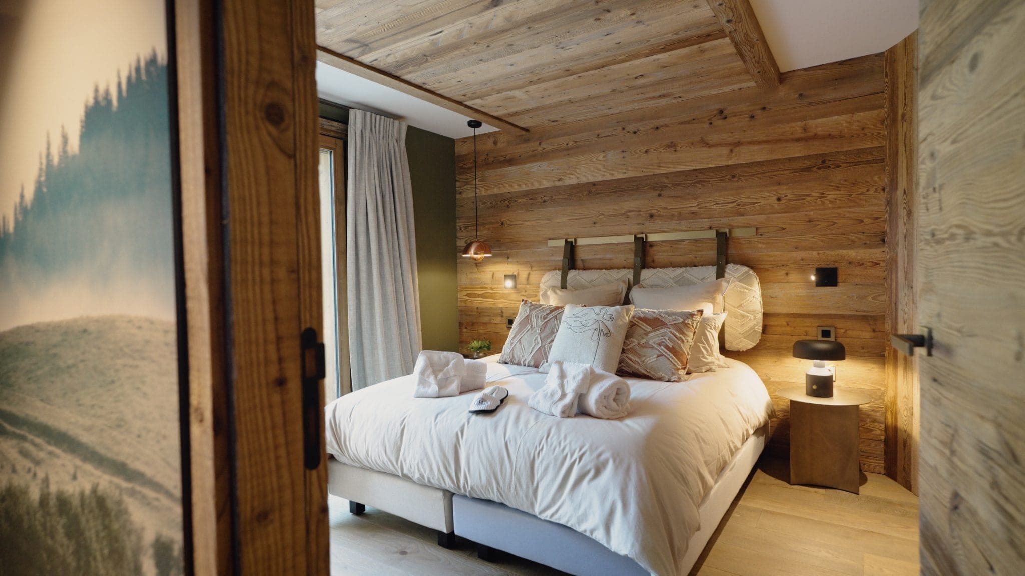 Cozy bedroom with wooden walls and ceiling, featuring a neatly made bed with plush pillows and towels. A bedside table with a lamp is beside the bed. Natural light streams in through a window with curtains, creating a warm and inviting atmosphere.