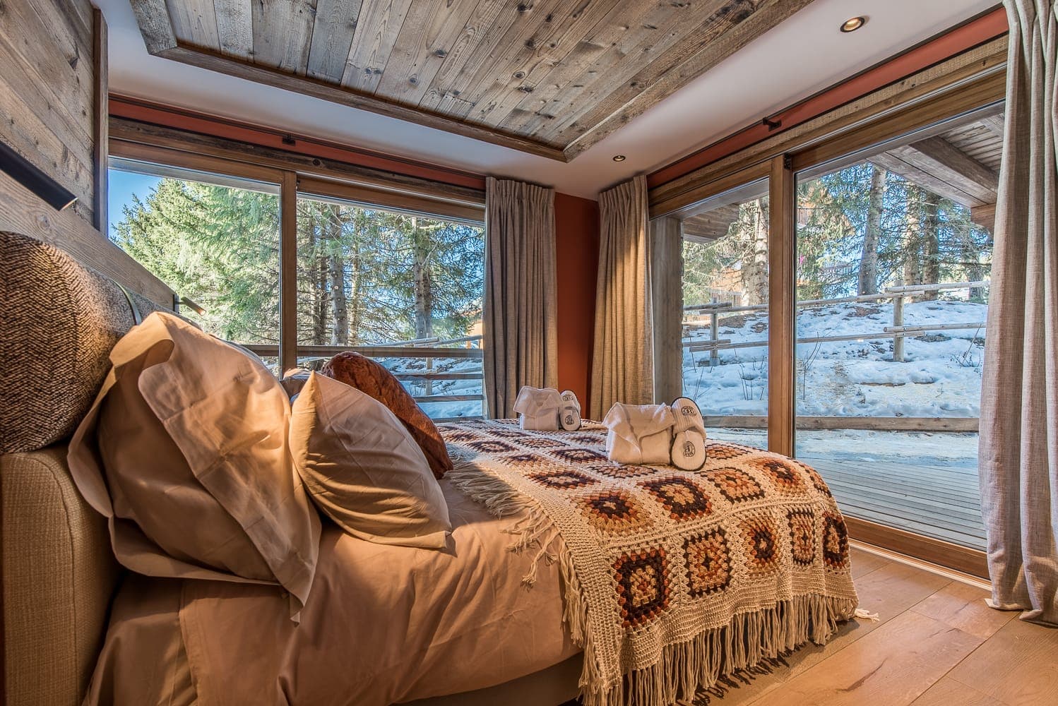 Cozy bedroom with a large bed adorned with patterned throw and pillows. Towels are neatly placed at the foot of the bed. Large windows provide a view of snowy trees and a wooden deck outside. Warm lighting and wood accents create a welcoming atmosphere.
