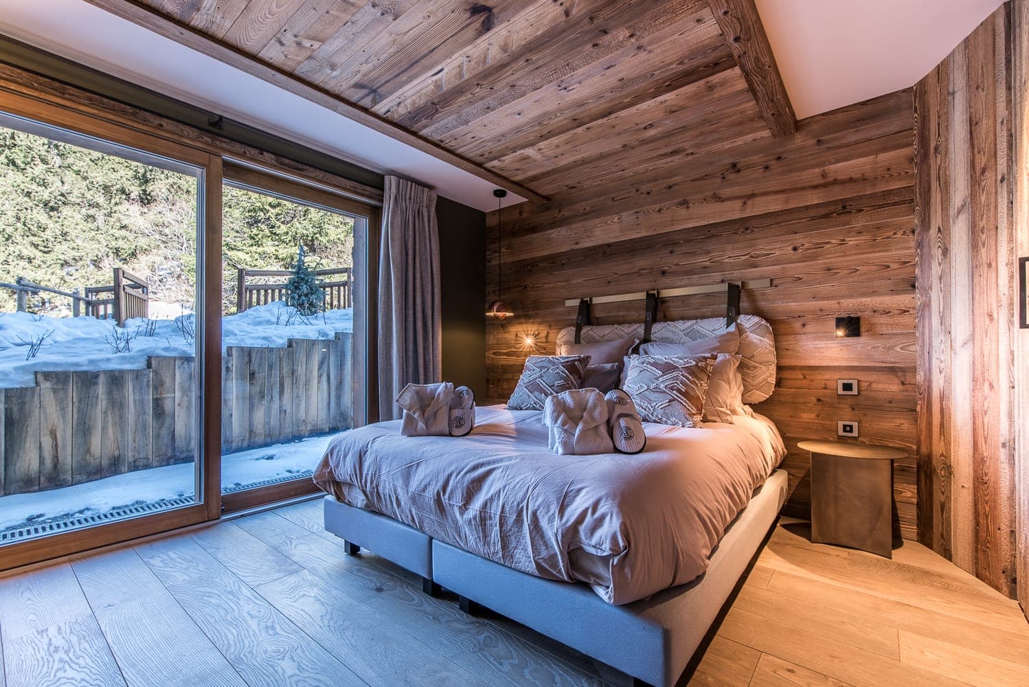 A cozy bedroom with a wooden interior, featuring a double bed adorned with pillows and towels. Large glass doors open to a snow-covered landscape outside, bringing in natural light. Dark curtains and wooden floors complement the rustic, warm ambiance.