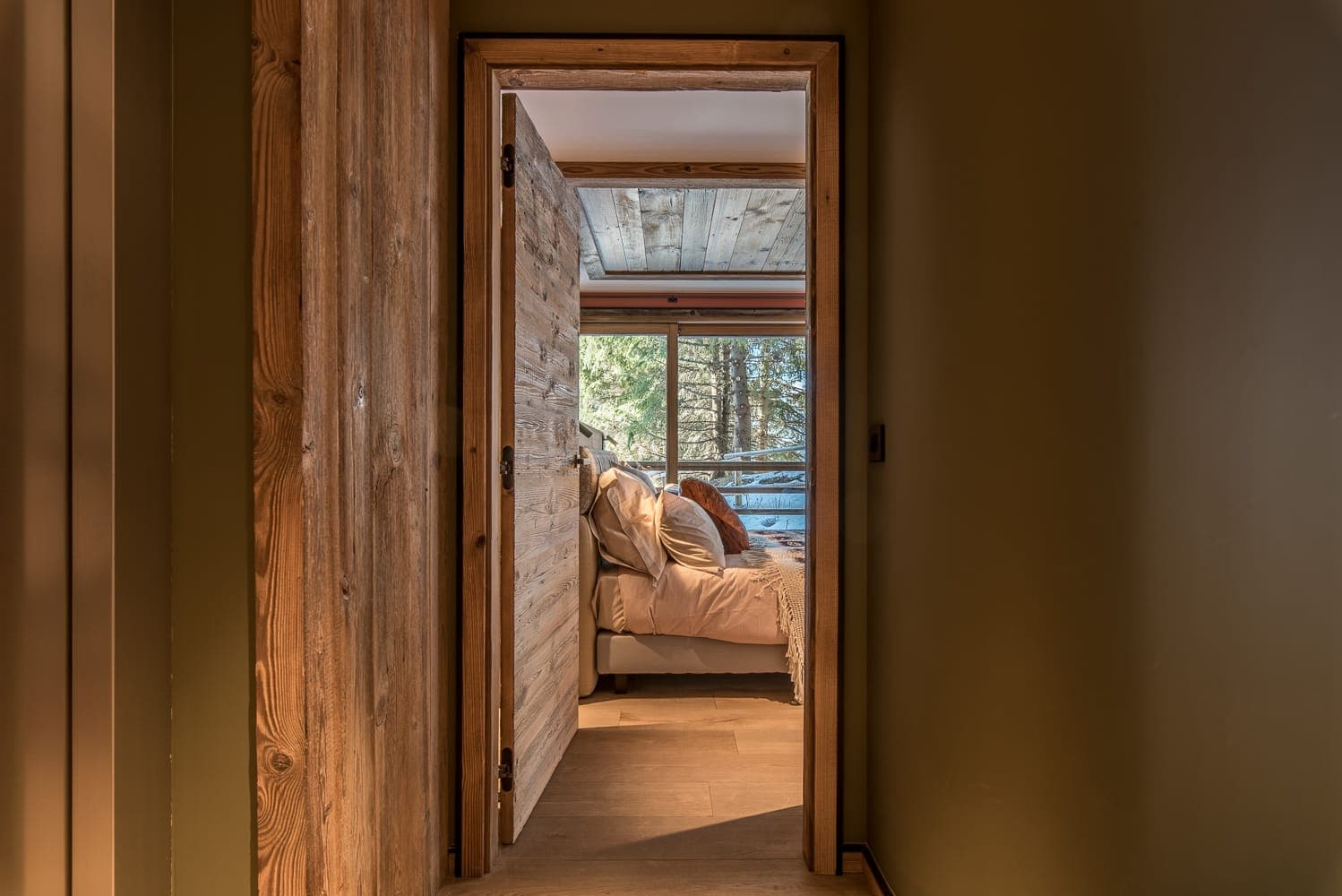 A cozy bedroom is visible through an open wooden door. The room features a neatly made bed with white linens and decorative pillows, a wooden ceiling, and a large window that lets in natural light, showcasing a view of trees outside.