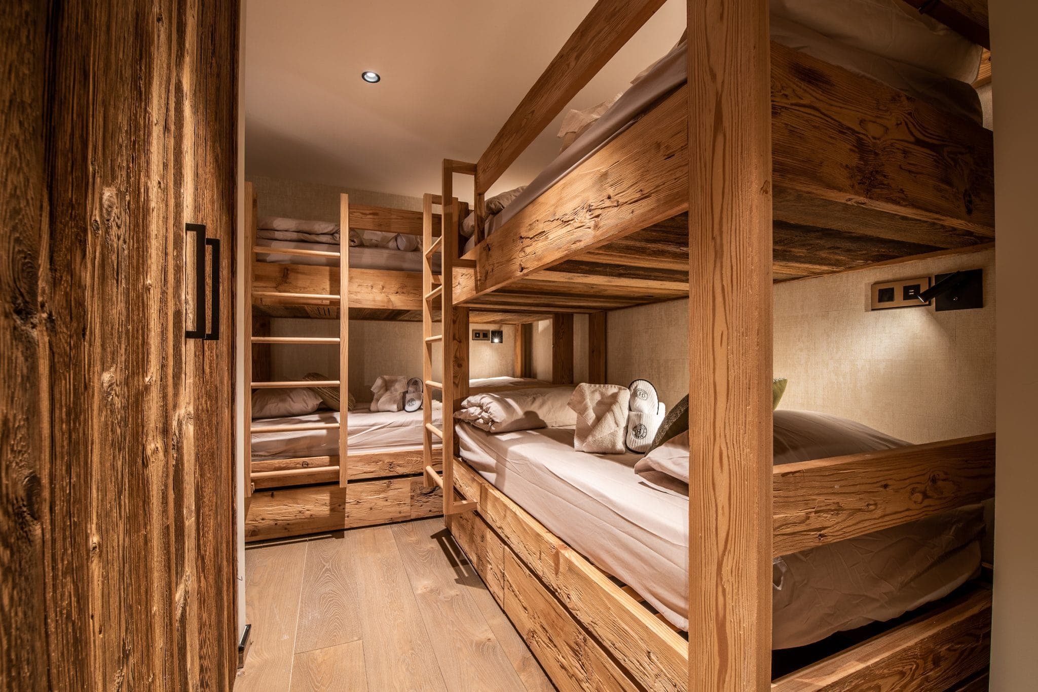 Wooden bunk beds with white bedding in a cozy, narrow room featuring warm lighting. The room's walls and floor are also wooden, creating a rustic and inviting atmosphere. A small window is visible on one side.