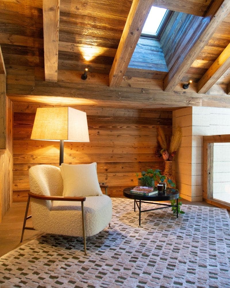 Cozy room with a wooden interior featuring a plush chair beside a lit floor lamp. A skylight illuminates the space, which includes a small round table with plants and books. A patterned rug covers the floor, complementing the rustic ambiance.
