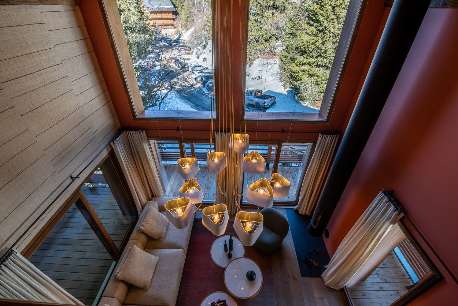 Aerial view of a cozy living room with a large, modern chandelier and beige furniture. Floor-to-ceiling windows reveal a snowy outdoor scene with trees and a parked car. Cream curtains frame the windows, adding warmth to the space.