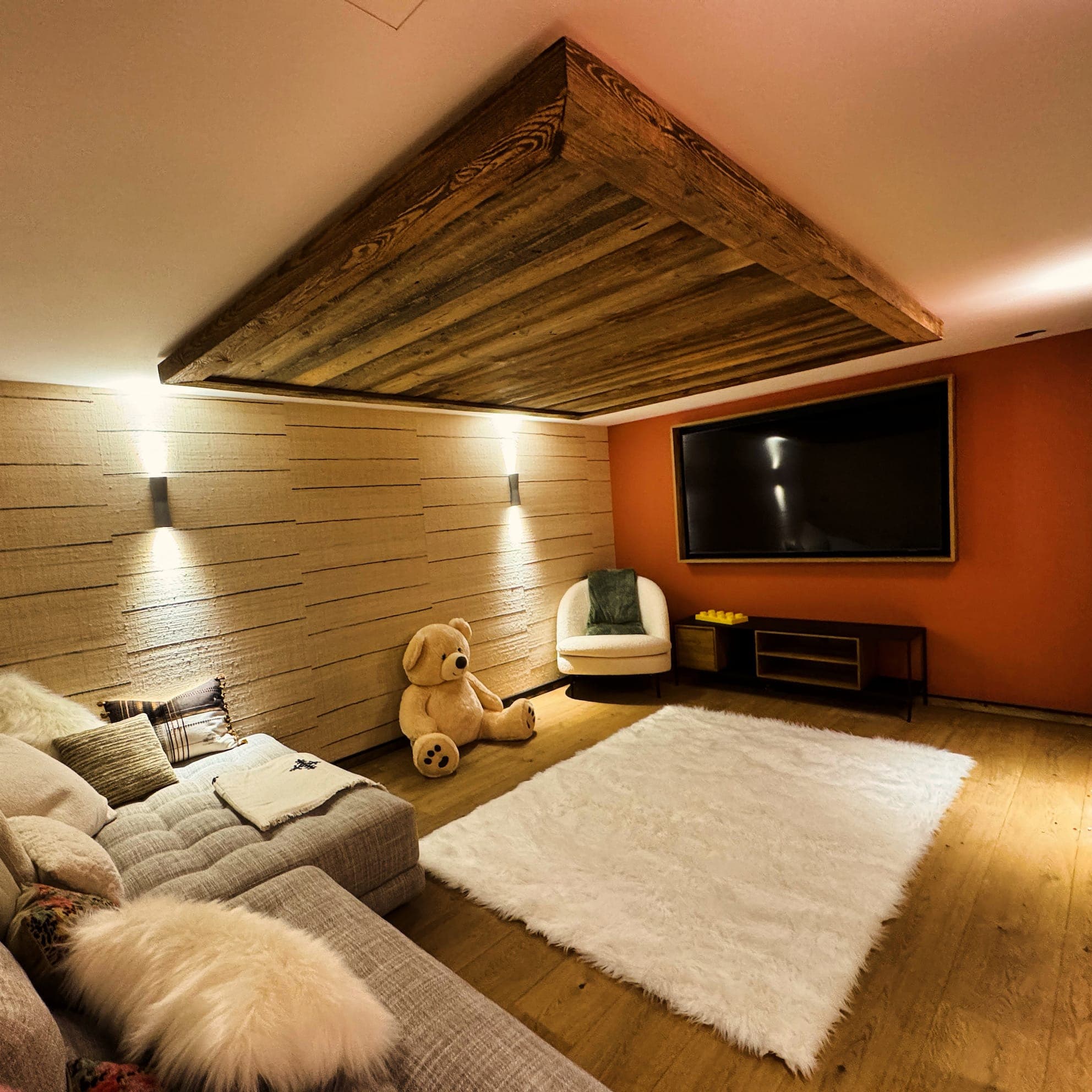 Cozy living room with a large TV on an orange accent wall, a textured wooden ceiling panel, and a plush white rug. A big teddy bear sits on the floor near a white armchair. The grey couch has fluffy cushions and a throw. Warm lighting enhances the ambience.