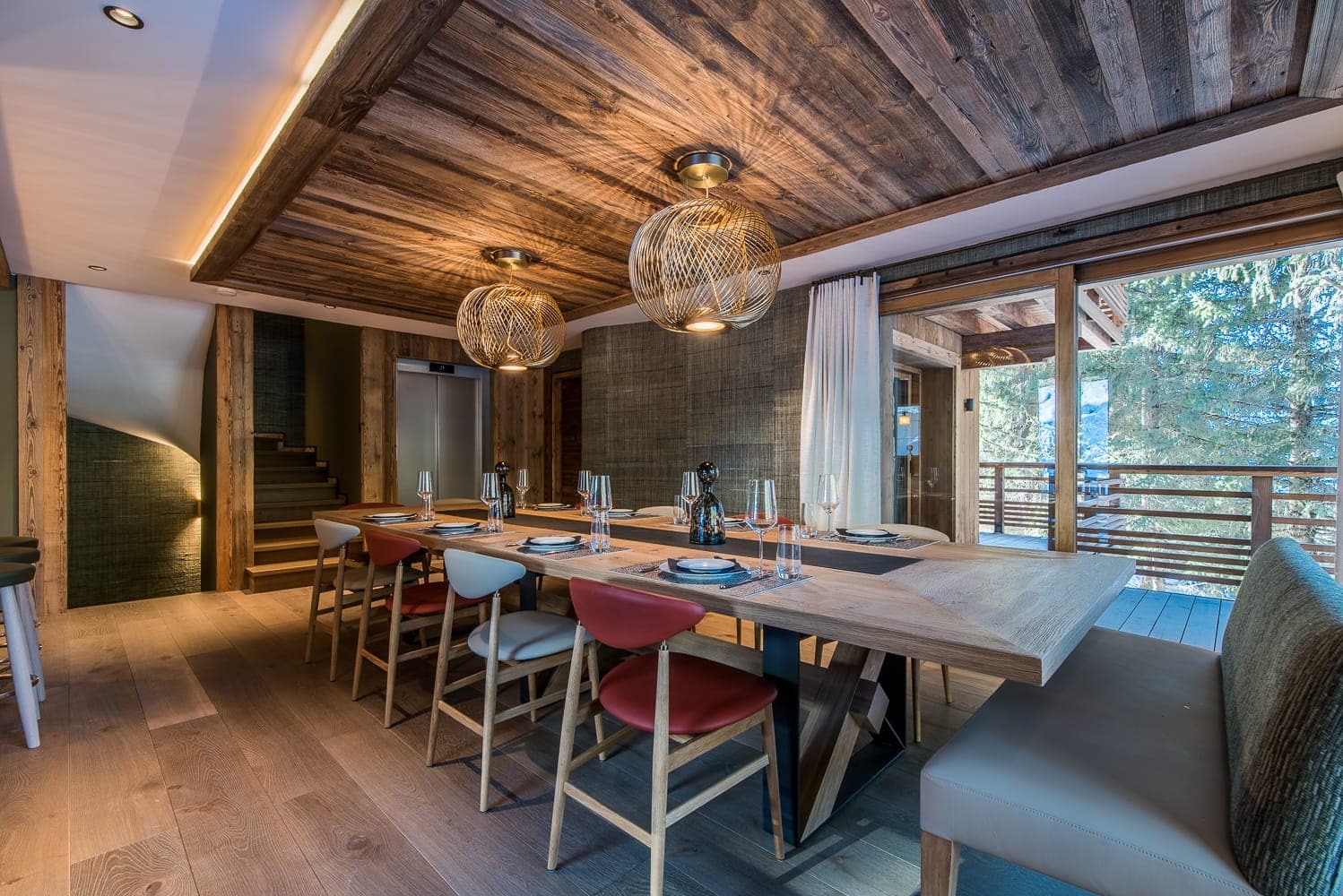 A modern dining area with a wooden table set for six, featuring colorful chairs and stylish pendant lights. The space has a rustic wooden ceiling and floor, with glass doors opening to a balcony with views of trees.