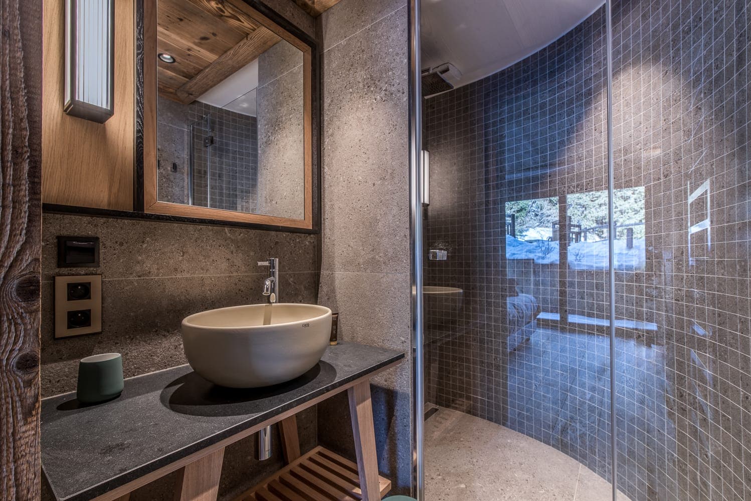 Modern bathroom with a round glass shower, gray tiled walls, and a wooden vanity. The sink is bowl-shaped beside a green cup. A large mirror reflects the well-lit, rustic decor, and a window provides natural light and a view of trees.