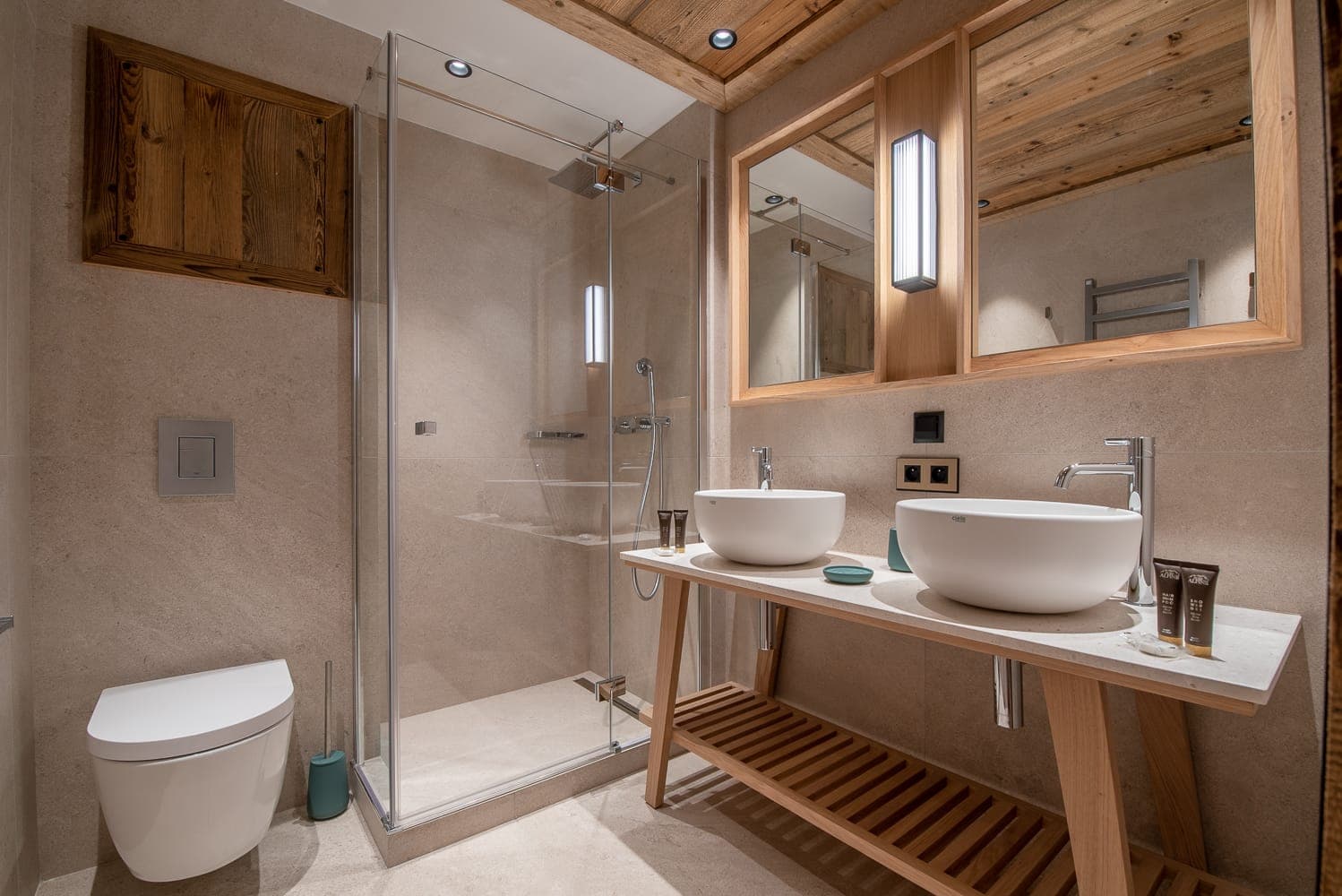 Modern bathroom with a glass-enclosed shower, dual round sinks on a wooden vanity, rectangular mirrors, and a wall-mounted toilet. The space features wood accents on the ceiling and mirrors, creating a warm, rustic feel.