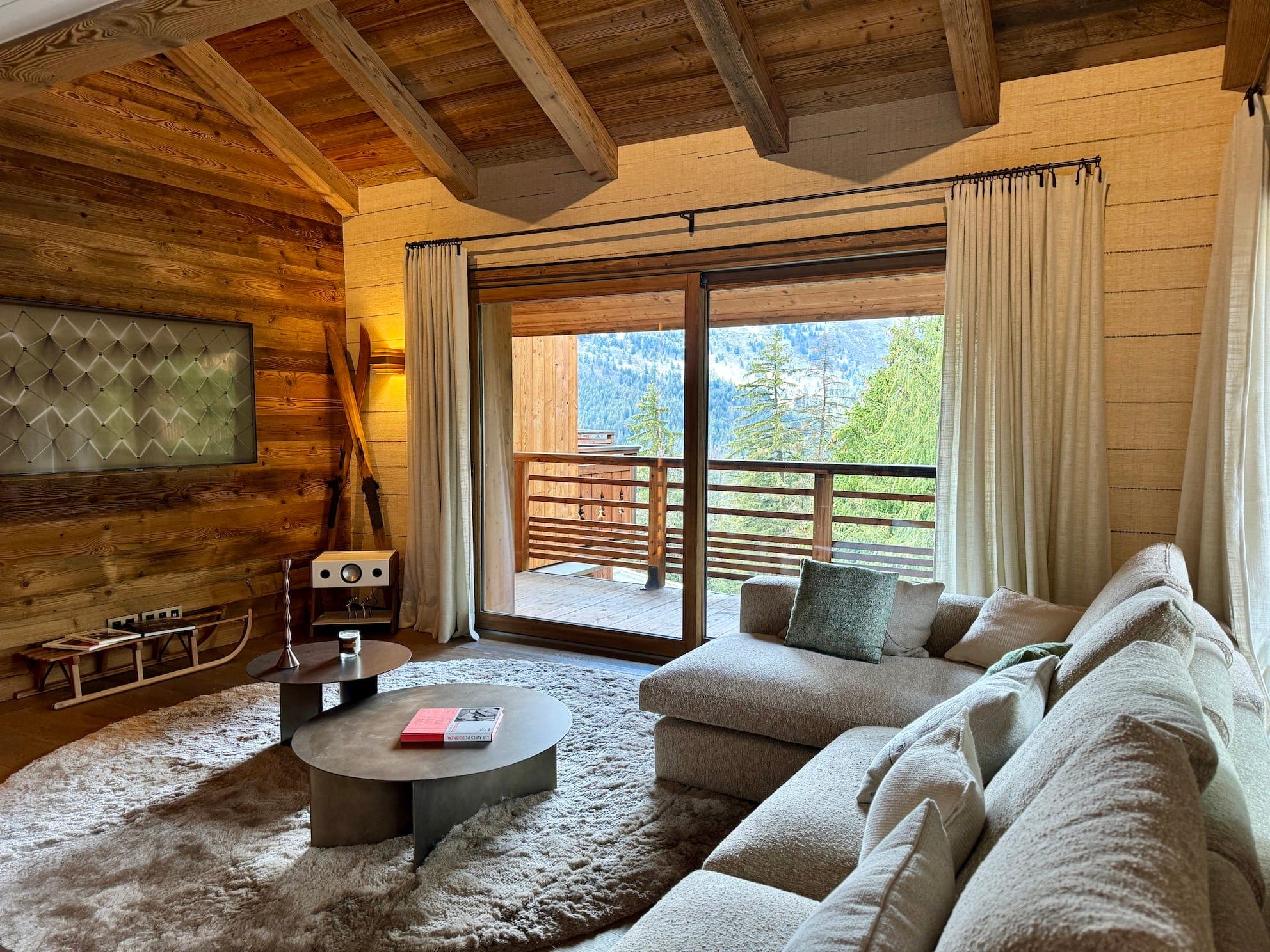 A cozy living room with a large beige sectional sofa and circular coffee table on a plush rug. Large windows with curtains open to a wooden balcony and scenic view of trees and mountains. Warm wood paneling adds a rustic touch.